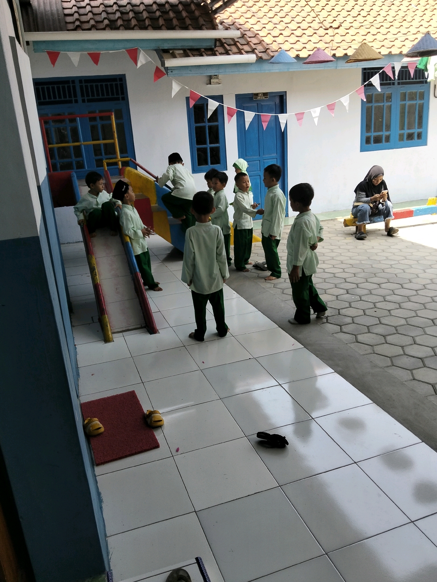 In Krajan Lor, Indonesia, a group of children in light green uniforms are gathered on a tiled courtyard in front of a building with a red-tiled roof. Some children are playing on a red, yellow, and blue slide. A woman in a dark hijab and jeans sits on a yellow bench, looking at her phone. The courtyard is bordered by a white-tiled area and a darker tiled area with a hexagonal pattern. Small red and white flags are strung across the front of the building, which has blue-framed windows. Two pairs of yellow children's sandals are visible on a red mat.
