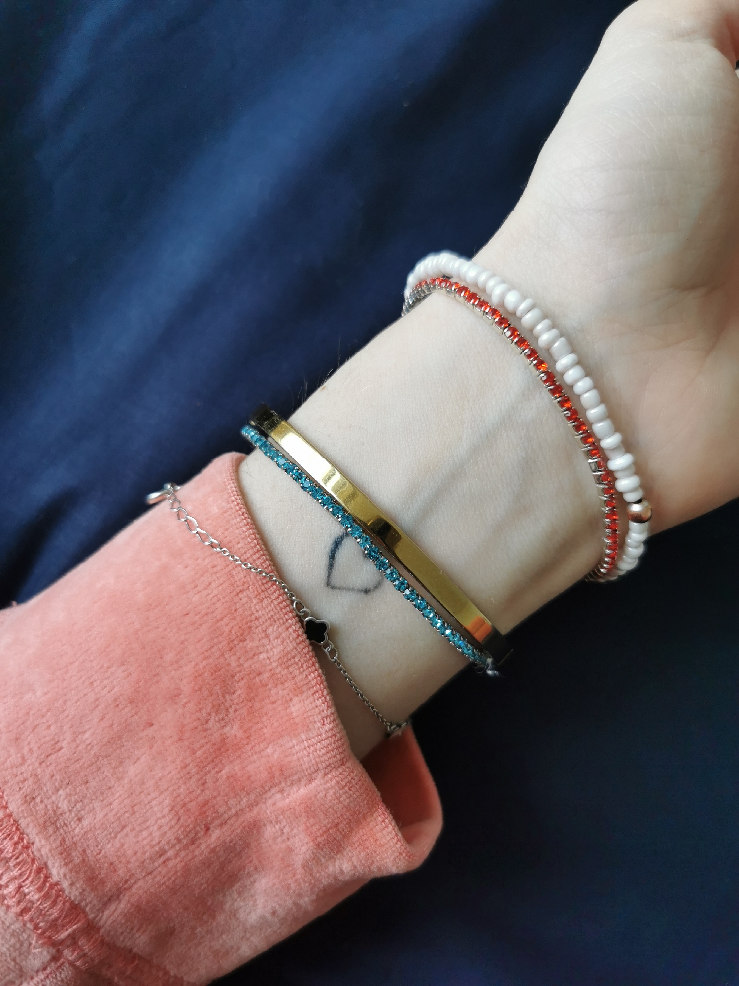 A close-up shot displays a person's wrist adorned with multiple bracelets. The wrist is positioned against a blurred background of dark blue fabric. The person is wearing a pink soft fabric top with visible ruching detail.
The bracelets consist of the following:
- A string of white beads with alternating red beads.
- A gold-toned, thin bangle.
- A bracelet featuring small blue stones set in a metal frame.
- A delicate silver-colored chain bracelet with a black clover-shaped charm.
A small tattoo of a heart is visible on the wrist.
The location is not identifiable from the image.