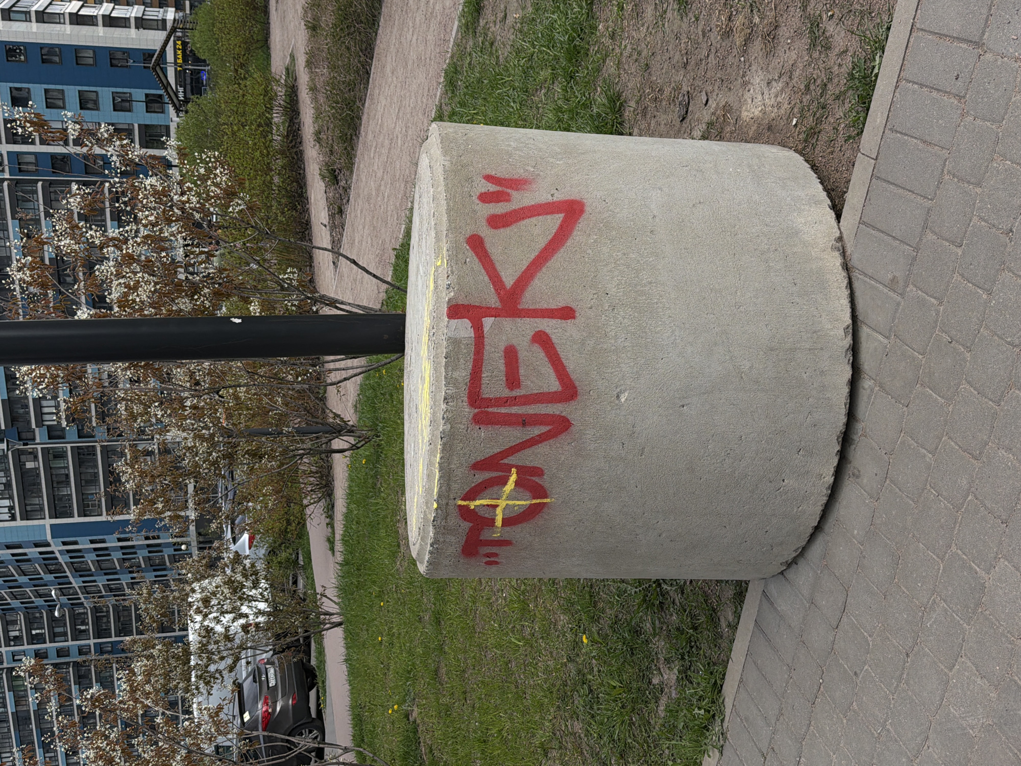 A cylindrical concrete structure is situated next to a gray brick pathway bordered by green grass and a low hedge. Red graffiti, potentially in Russian, is spray-painted vertically on the side of the cylinder. A black metal post is positioned adjacent to the cylinder, with a flowering tree or bush growing nearby. In the background, multi-story residential buildings are visible. The overall environment suggests an urban park setting in Murino, Russia.