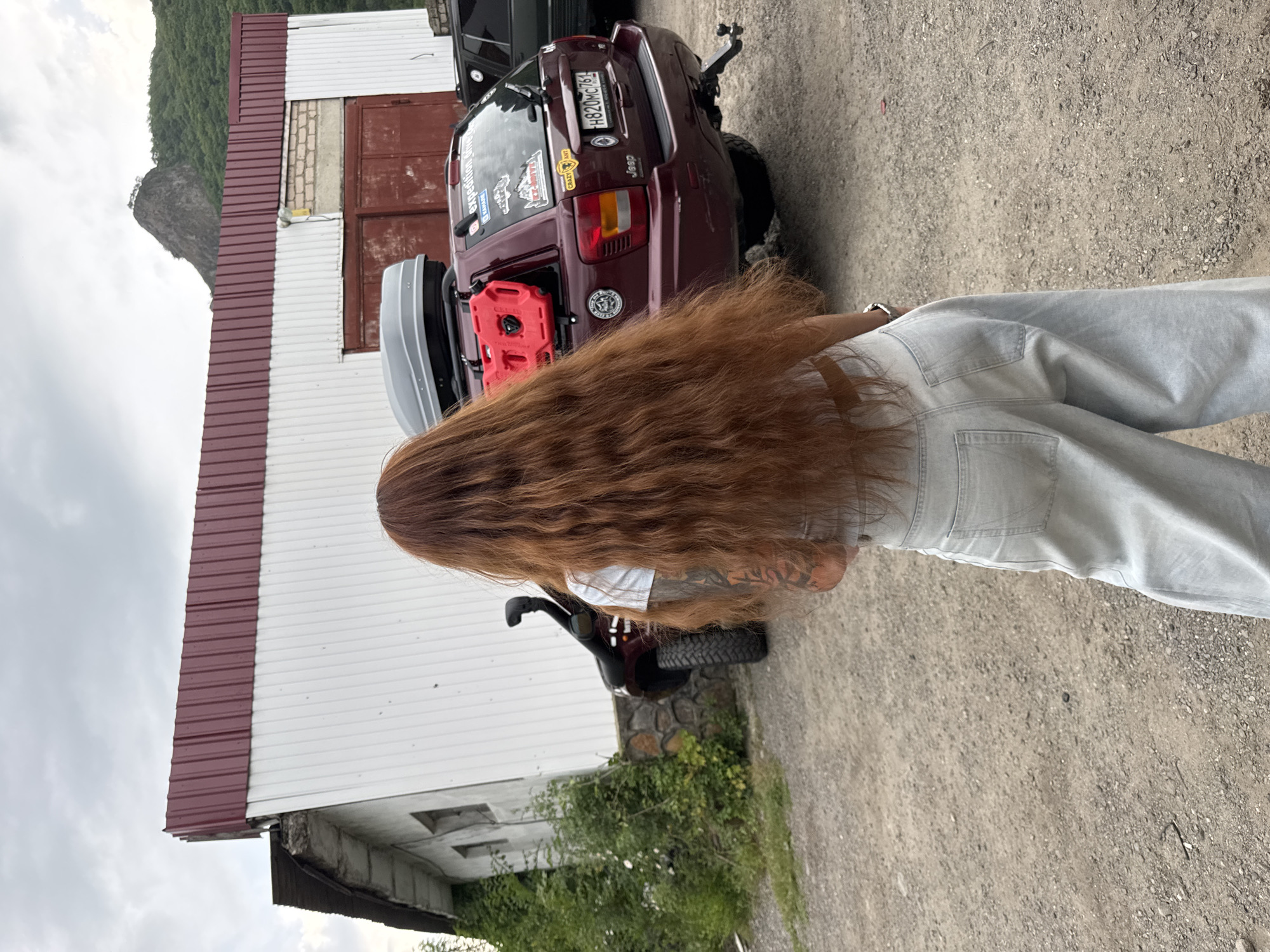 A full-body shot from behind shows a woman with long, auburn hair standing on a gravel road. She is wearing a white crop top and light blue, wide-leg jeans. A dark red Jeep Grand Cherokee is parked behind her. The Jeep has a roof-top cargo carrier, a red jerry can mounted on the rear passenger side, and several stickers on the back window, including one that says "Jeep" and another with a license plate reading "H820MC761". A snorkel is attached to the driver's side of the Jeep. In the background, there is a long, two-story building with a white corrugated metal siding and a dark red metal roof. A mountain is visible in the distance. The scene appears to be in the city of Karachayevsk, Russia, given the architectural style and the landscape.