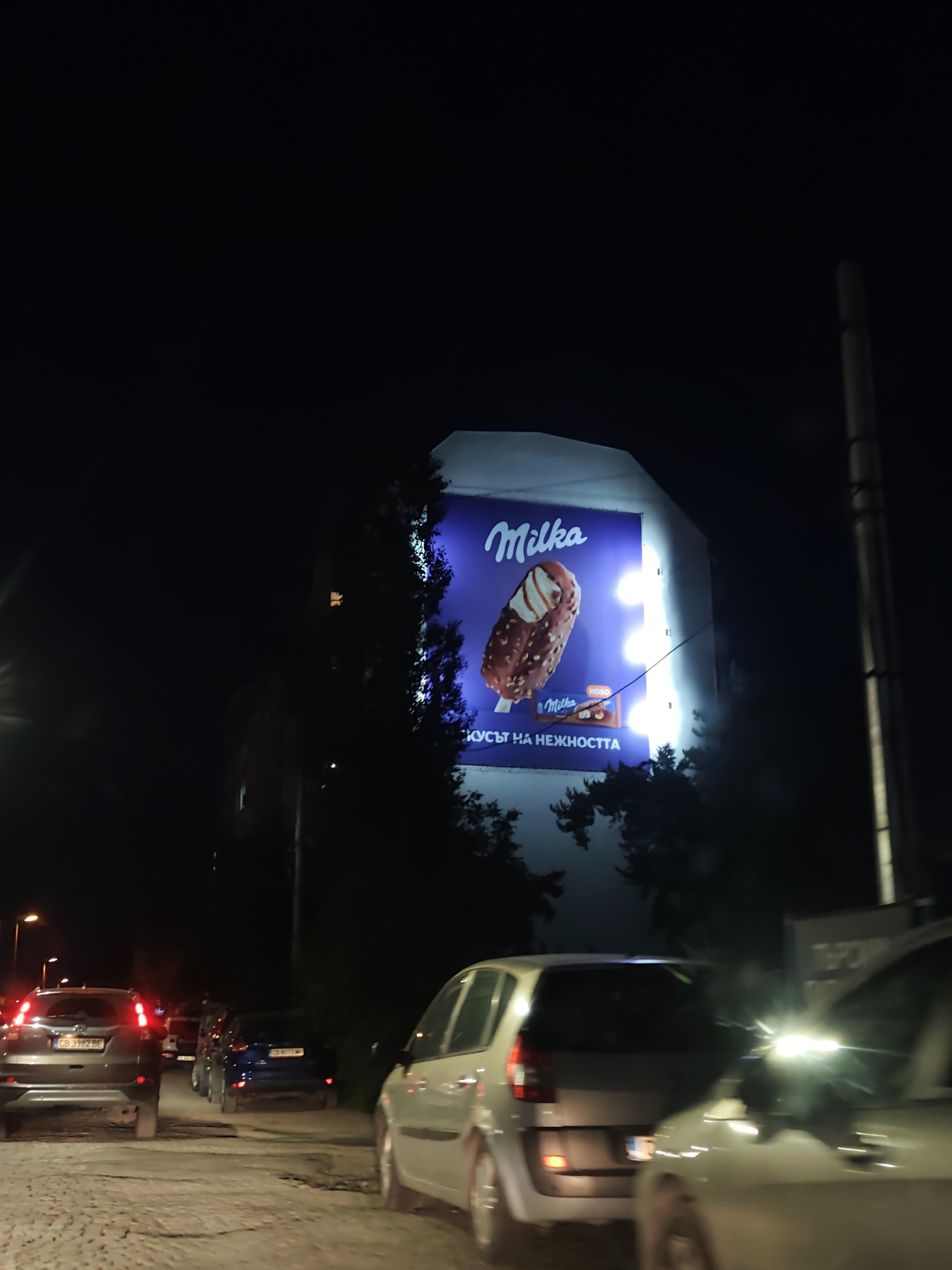 At night, a Milka billboard advertising ice cream is illuminated in Sofia, Bulgaria. The billboard, featuring the Milka logo and an image of a chocolate-covered ice cream bar, is mounted on a building. The scene is taken from a street with multiple cars, some with their headlights and taillights visible, indicating slow-moving traffic. Vegetation is visible on both sides of the billboard and the street. The road appears to be paved with cobblestones. A utility pole is seen to the right of the billboard.