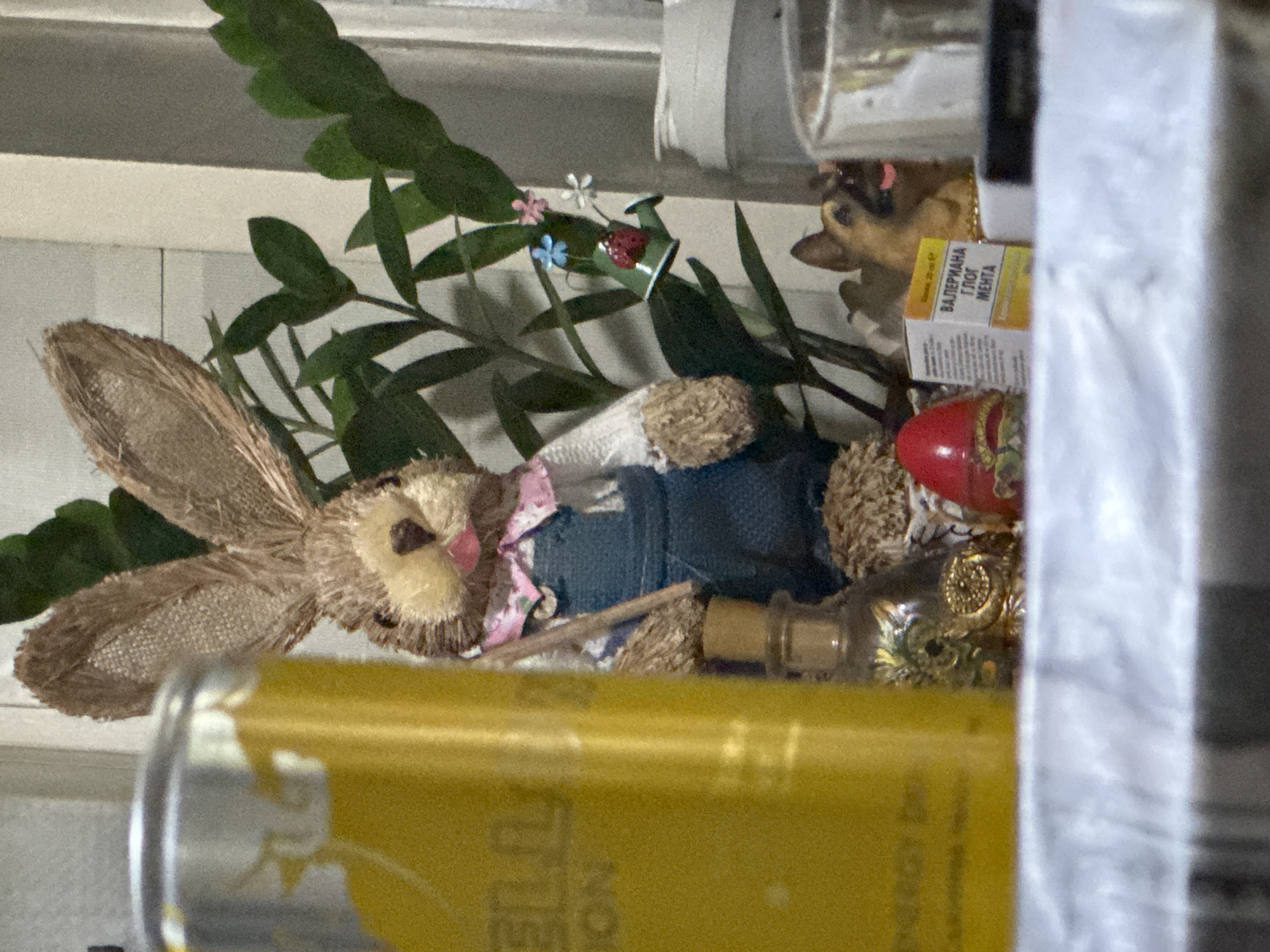 The image depicts a still life arrangement on a surface covered with a white and gray checkered cloth. In the foreground, a bright yellow can with partially visible text is positioned on the left side. To the right of the can, a small decorative bunny figure is visible, wearing a pink floral bandana and blue overalls. The bunny holds a small stick. Behind the bunny, a potted plant with dark green leaves is present. Among the leaves, a tiny green watering can with a ladybug and small flowers is attached. Further to the right, a small figurine of a German Shepherd dog is placed. In front of the dog, a small box with text in Cyrillic script is visible, with the words "ВАЛЕРИАНА ГЛОГ МЕНТА" clearly readable. A red, egg-shaped object is also part of the arrangement, situated to the left of the dog figurine. The background consists of a light-colored surface, possibly a wall with horizontal paneling. The lighting suggests an indoor setting. There are no people visible in the image. The scene does not provide any specific indicators of its location within the city of Sofia, Bulgaria.