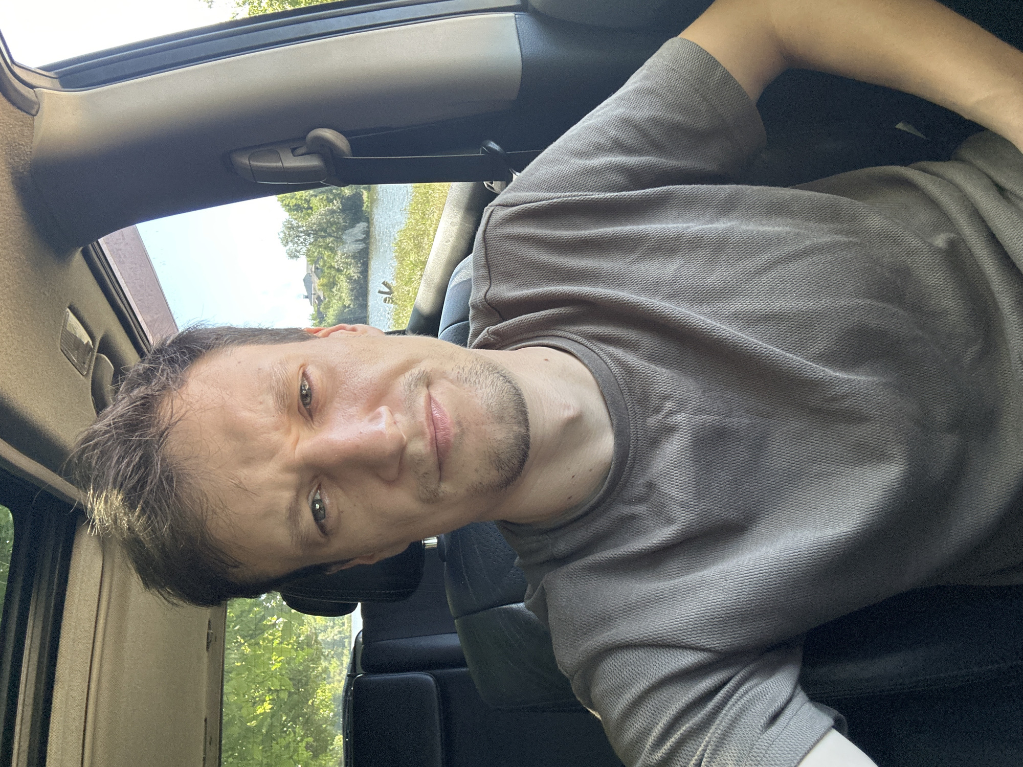 A man is sitting in the driver's seat of a vehicle, taking a selfie. The man has brown hair and a short beard and is wearing a grey t-shirt. He is looking directly at the camera with a slight frown. Outside the car window, trees, a body of water, and some buildings can be seen. The image appears to have been taken during the daytime. The setting is consistent with Makiv, Ukraine, a village located in the Vinnytsia Oblast.