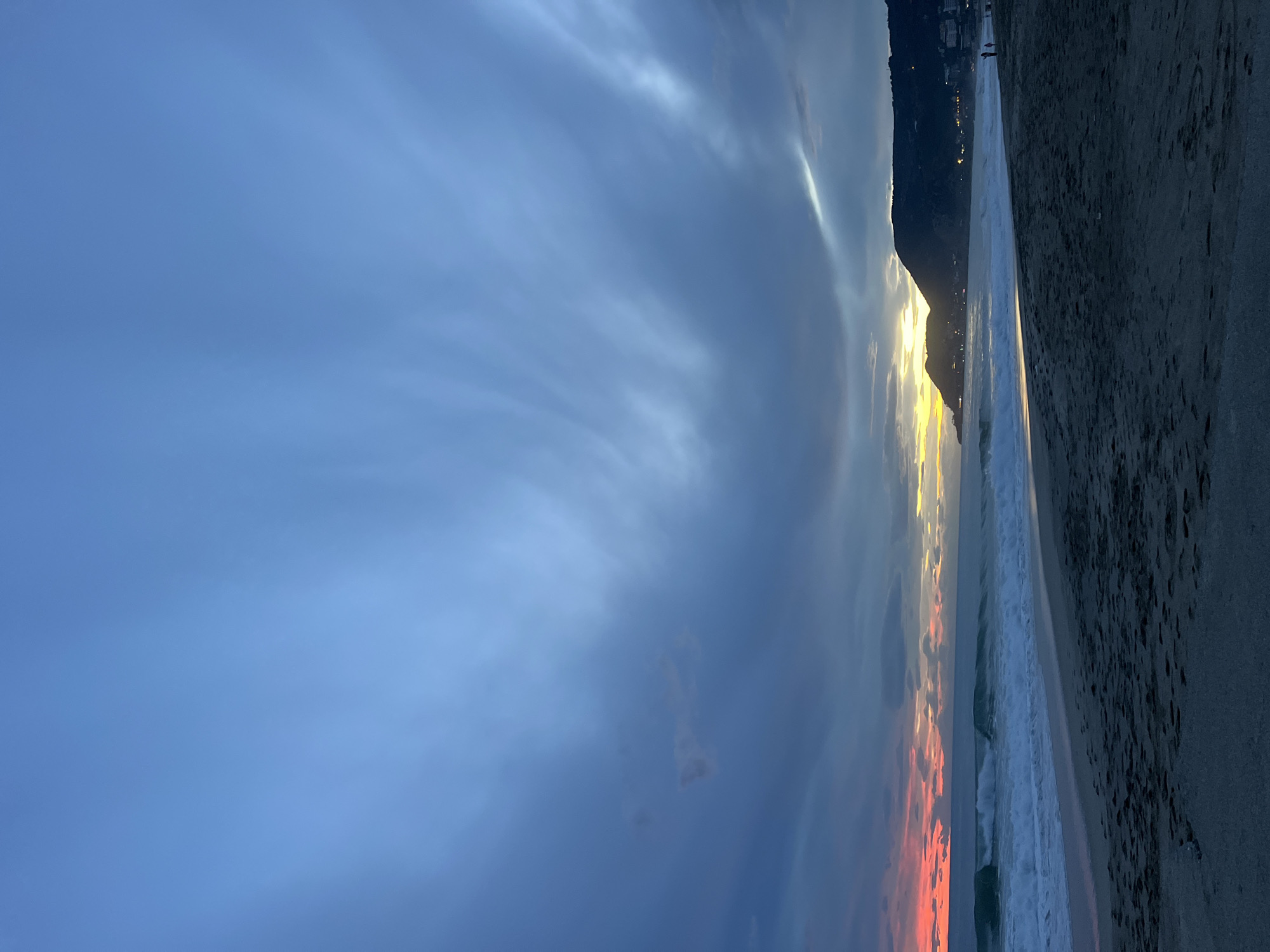 The image shows a beach scene at sunset in Alanya, Turkey. A dark, sandy beach occupies the right side of the image. Waves are breaking on the shore. A dark mountain silhouette lines the coast in the center of the image, with some scattered lights indicating a settlement. The setting sun casts a golden glow behind the mountain, and some orange colors are reflected in the clouds near the horizon. The sky is predominantly blue, with streaks of clouds. Two small figures are visible in the distance at the edge of the beach and the mountain.