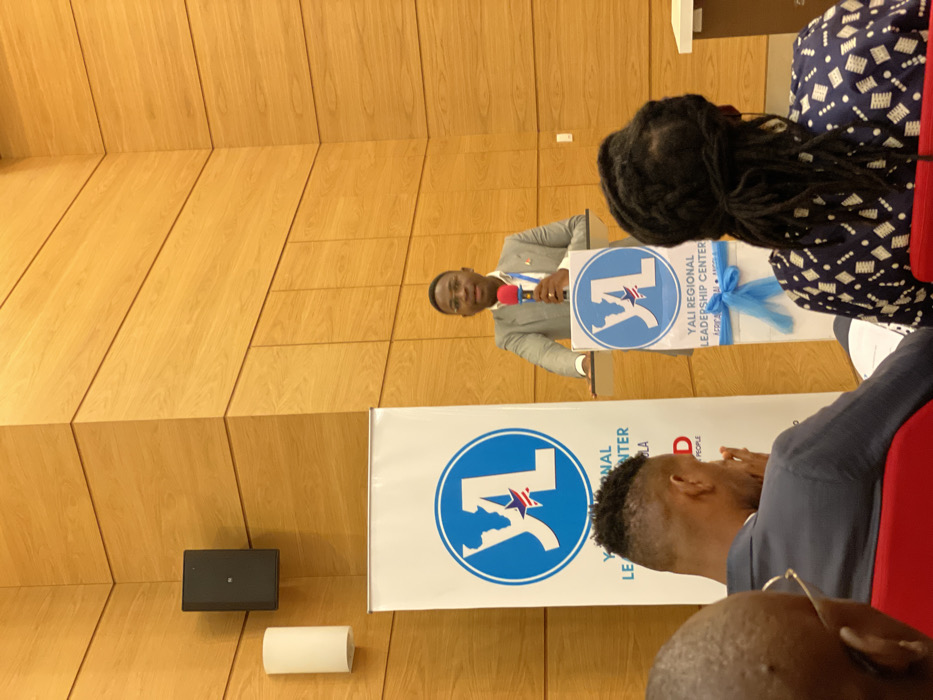 The image captures an indoor event, likely a presentation or conference, seen from an audience perspective. A man, dressed in a light-colored suit, stands behind a clear podium on the left side of the frame, speaking into a red microphone. He is addressing an unseen audience to his right.
The podium features a banner with a blue circular logo displaying a white stylized "YL" and a star. Text on this banner clearly reads "YALI REGIONAL LEADERSHIP CENTER" and "ACRP LUANDA ANGOLA". A light blue decorative ribbon is tied around the podium. Behind the podium, a larger, floor-standing banner with the same YALI logo is partially visible, also bearing the text "YALI REGIONAL LEADERSHIP CENTER" and the word "PEOPLE".
The background wall is covered with light-colored wooden panels arranged in diagonal sections. A black speaker box is mounted on this wall to the left of the speaker, and a cylindrical white object rests on the floor below it.
In the foreground, on the right side, parts of several audience members are visible from behind. One person has long dreadlocks and wears a dark patterned top. Another person is seen from the back, sporting a faded haircut and wearing a dark suit jacket. A red seat or chair back is also visible in the lower right corner. The event appears to be a formal gathering, possibly a leadership development program, taking place in Luanda, Angola, as indicated by the visible text.