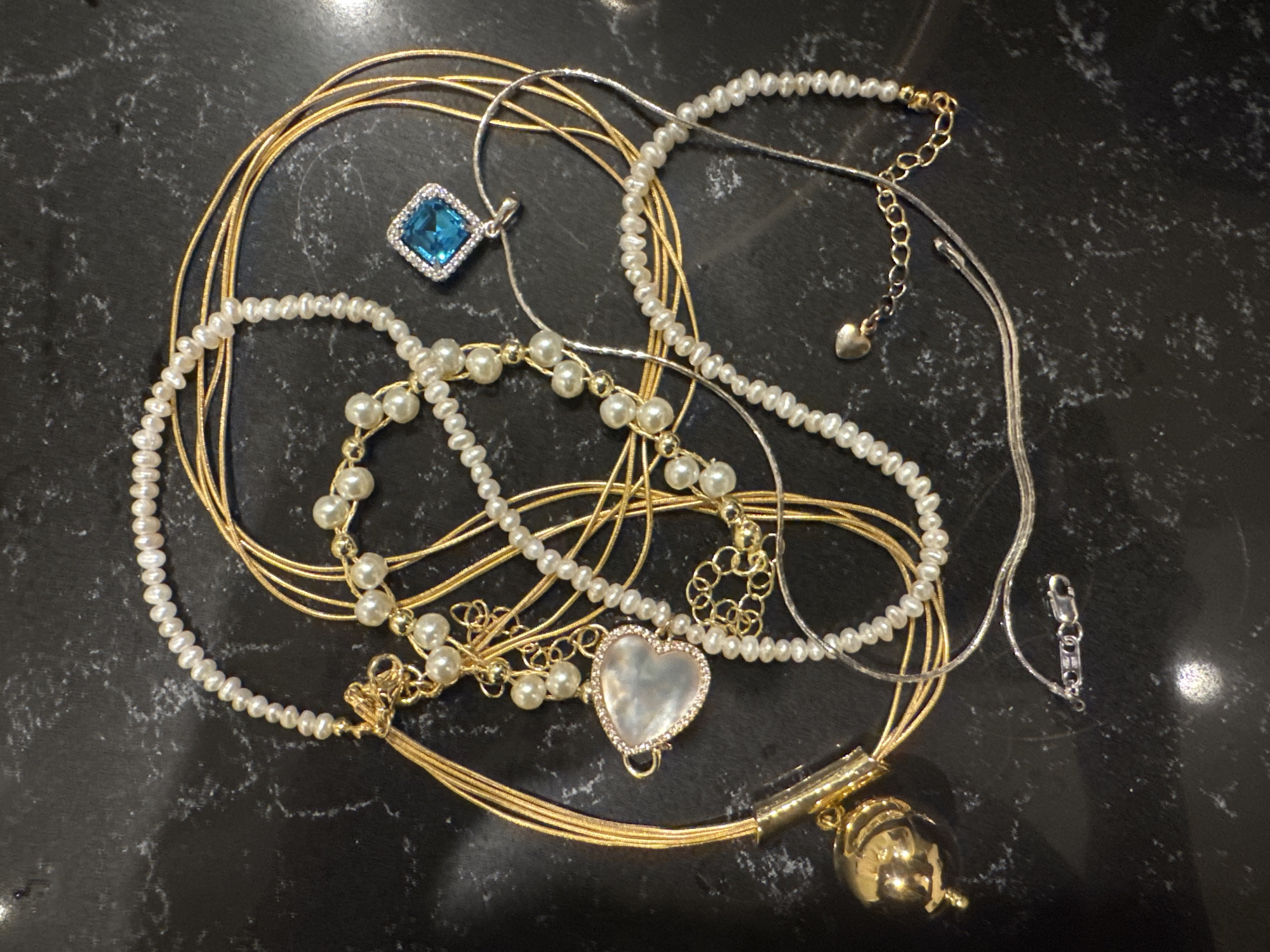 The image shows an assortment of jewelry items scattered on a dark, possibly marble, surface. The jewelry includes gold-colored necklaces, strands of pearls, and silver-colored necklaces. A blue square-shaped pendant with a silver-colored setting is visible. There is also a heart-shaped pendant with a pearlescent center. A gold-colored pendant with an ornate, rounded design is also present. The items are intertwined and overlapping.