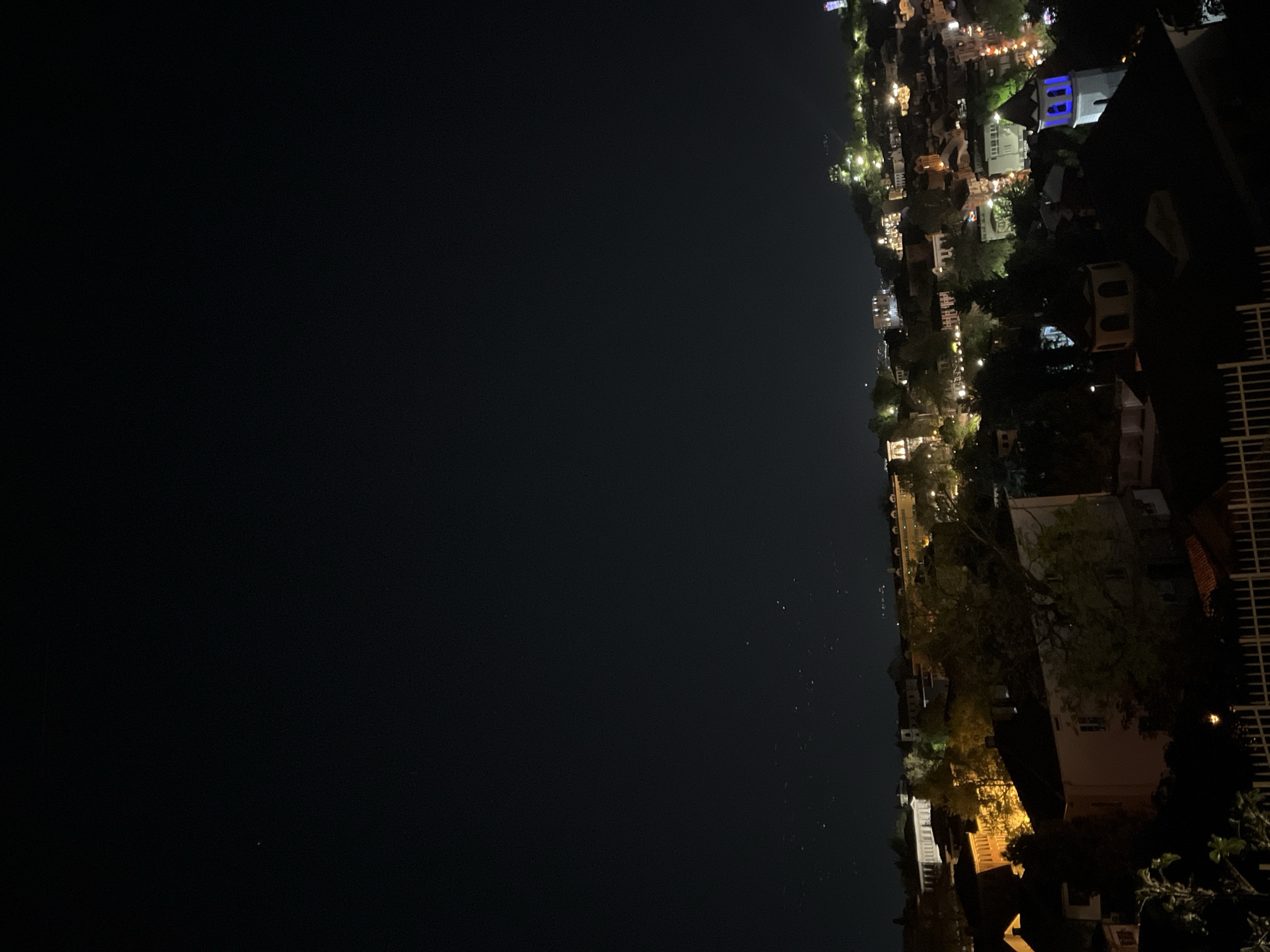 An eye-level, nighttime shot looking out over the city of Plovdiv, Bulgaria. The photo is oriented sideways, with the dark sky and body of water on the left and the illuminated city on the right. The city appears to be situated along the water's edge, with lights from buildings and streets visible. The city is dense with structures, with some recognizable shapes of buildings and trees interspersed with the lights. In the foreground, there's a structure with what appears to be a fence, possibly a balcony or rooftop. The sky is dark, with a few indistinct specks that might be stars.