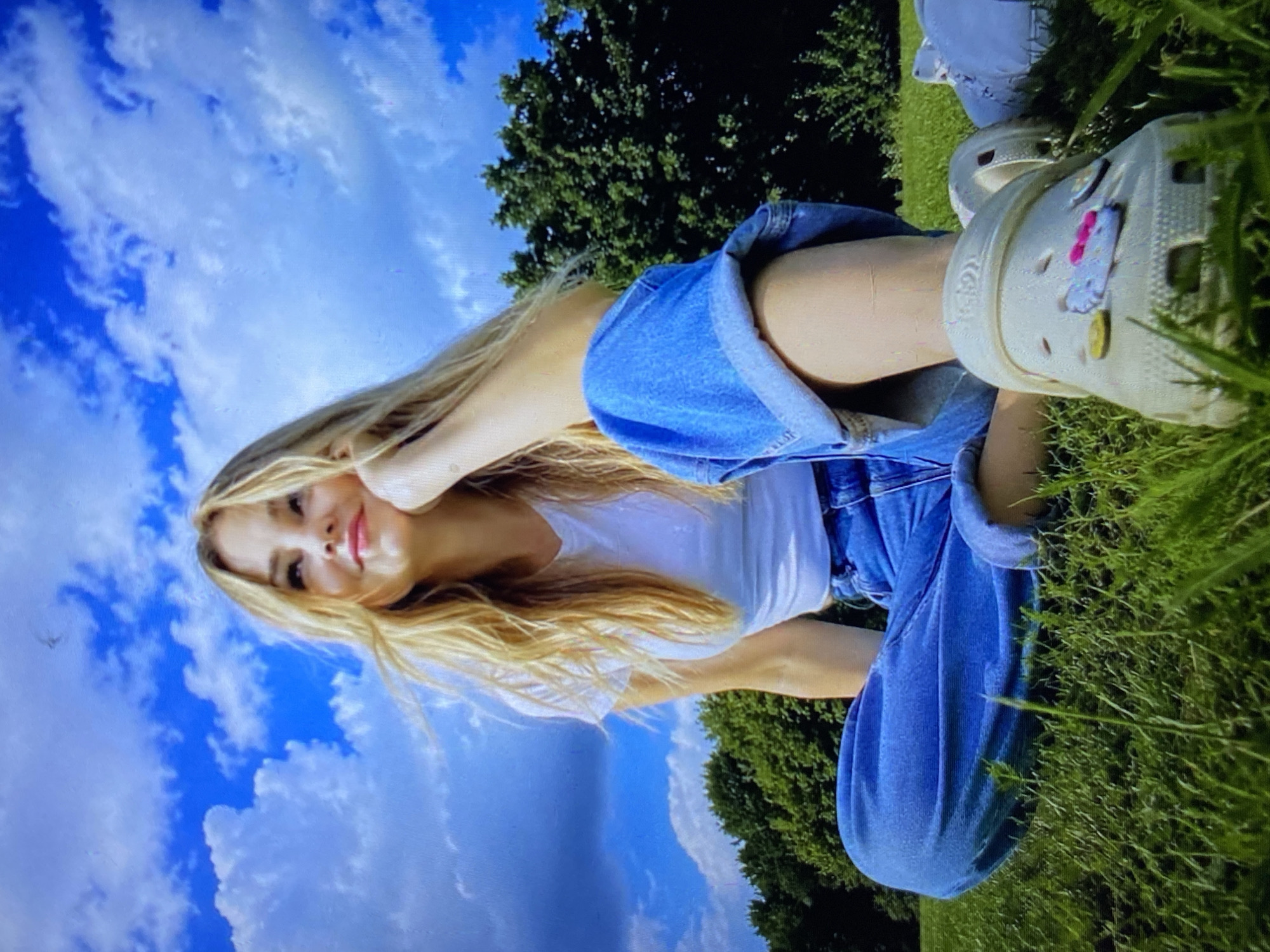 The image depicts a young woman with blonde hair sitting on a grassy lawn, looking up at a bright blue sky with scattered white clouds. She is wearing a white t-shirt and loose-fitting blue jeans, and her feet are adorned with light-colored clogs decorated with colorful charms. The background features lush green trees. There is no indication of the location being the city of Luh, Ukraine.