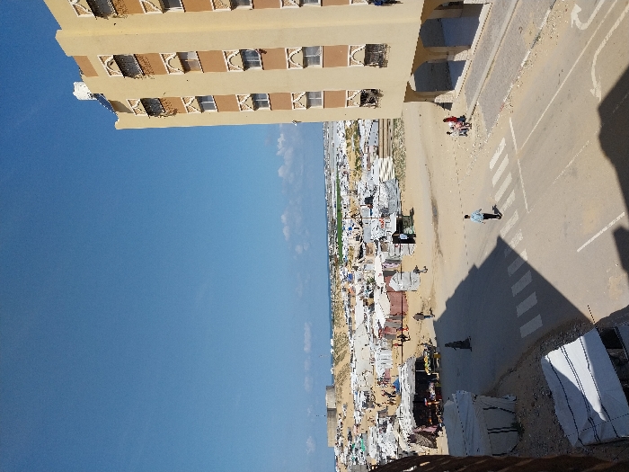 An elevated view captures a street scene in Khan Yunis, Palestinian Territory, under a mostly clear blue sky with sparse clouds near the horizon.
On the right, a multi-story, light yellow-orange building with brown architectural accents around its windows and dark grates occupies the frame. A white water tank and solar panels are visible on its roof. The building's ground floor features an arched arcade. Three individuals, two adults and one child, stand on the unpaved sidewalk adjacent to this building.
A paved road with a white pedestrian crosswalk and a white directional arrow traverses the mid-ground. A male individual in a light blue shirt and dark trousers walks from right to left across this crosswalk. Shadows, cast from an unseen structure, extend across the road and the lower left section of the frame.
To the left of the road, a vast and dense encampment of temporary shelters, constructed from tarpaulins, fabric, and corrugated metal, extends towards the horizon. Within this camp, multiple individuals are observed walking, and several makeshift market stalls displaying goods are visible under awnings. Further in the background, undulating sandy terrain leads to a prominent circular concrete structure on a rise, with the Mediterranean Sea visible beyond on the distant horizon. The ground in the camp and roadside areas is predominantly unpaved dirt and sand.