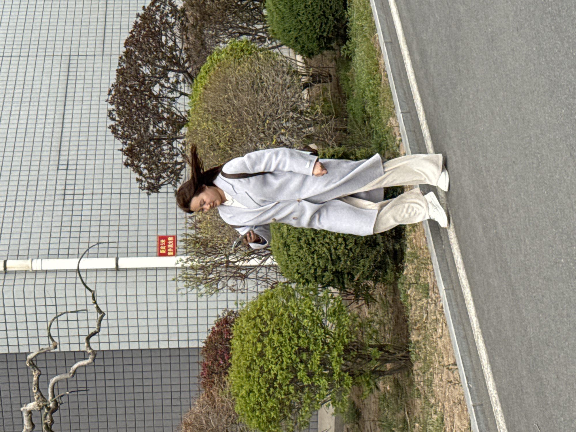 A woman is captured walking outdoors, engrossed in an object held in her hands, likely a phone, as she looks downwards. She is dressed in a light grey long coat, cream or light-colored wide-leg trousers, and white shoes. Her long, dark hair flows over her shoulders.
The setting is an outdoor area alongside a building and a road. To the woman's left, a verdant landscape features a variety of bushes and trees; some display vibrant green leaves, suggesting spring, while others are still bare or have reddish-brown foliage. Behind this greenery, a large, modern building with a distinct light-grey, grid-patterned facade dominates the background. To the woman's right, a grey paved road or path with a white curb runs along the length of the scene.
The lighting indicates daytime, appearing bright but diffused, possibly an overcast day, as there are no harsh shadows. A notable detail is a red sign attached to the building. The Chinese text on the sign reads: "距5米" (jù 5 mǐ), which translates to "5 meters away," and "室外消火栓" (shìwài xiāohuǒshuān), meaning "outdoor fire hydrant." The scene presents a casual, everyday moment in an urban or suburban outdoor environment.