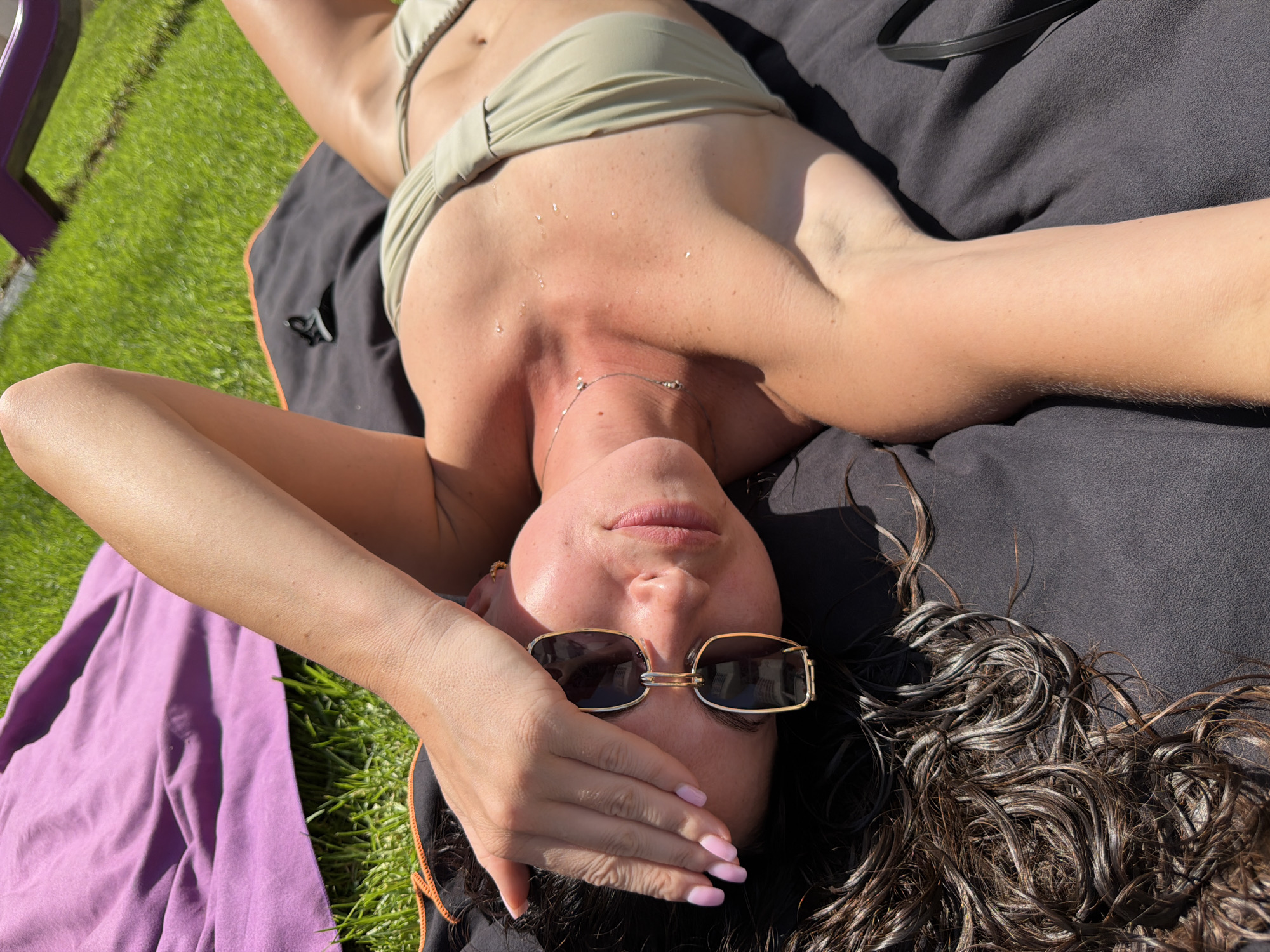 In Salou, Spain, a woman lies on her back on a dark towel, her body partially covered by a purple blanket. She wears a light green bikini, and sunglasses rest on her face, partially obscured by her hand, which is raised to her forehead. Her wet, dark, curly hair is spread out around her head. The background shows a green lawn and a hint of purple furniture. The scene suggests a sunny day outdoors, likely in a recreational area.