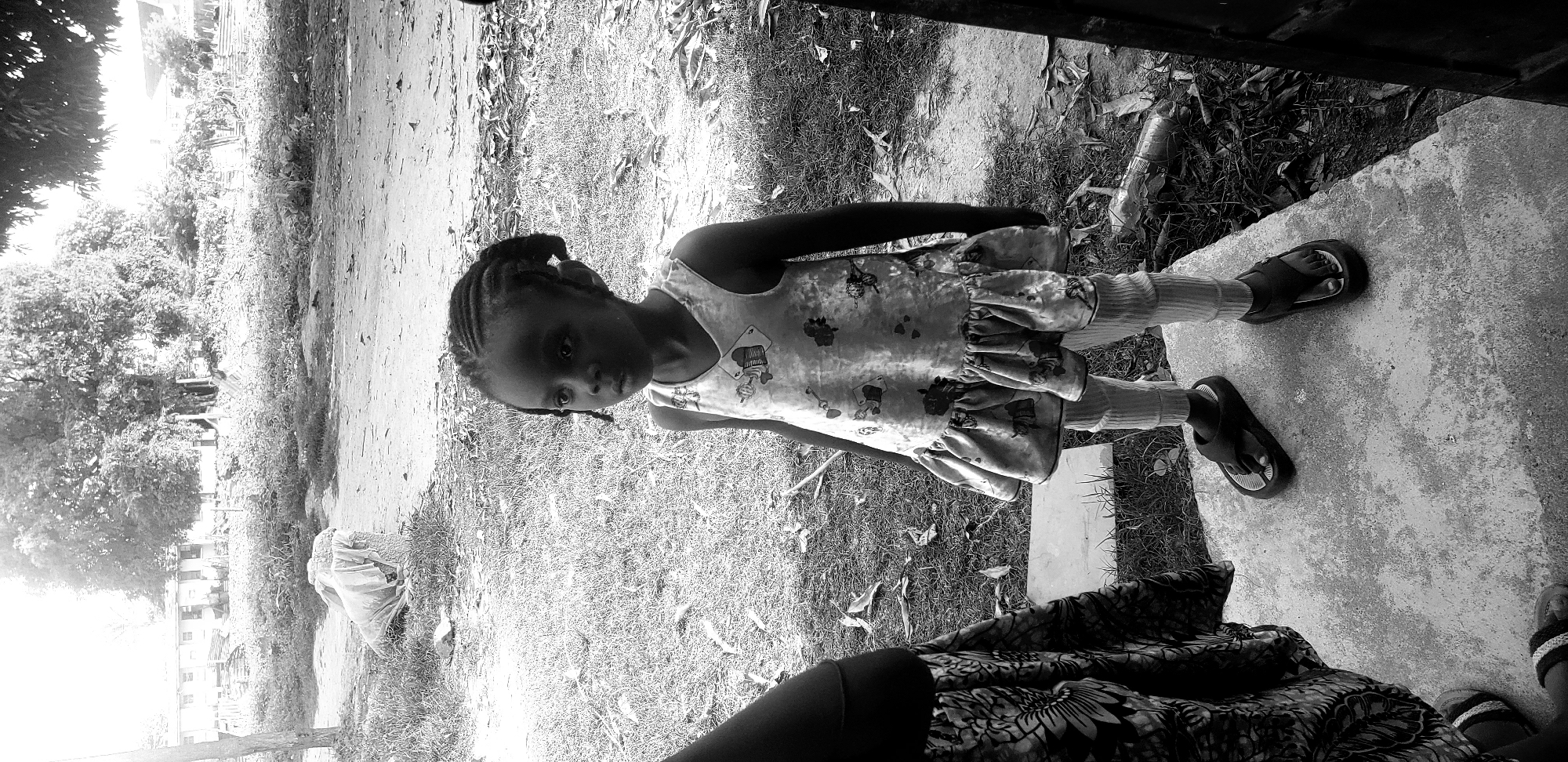 The image, captured in Narayi, Nigeria, shows a young Black girl standing on a concrete surface next to a grassy area. The girl is wearing a patterned dress with ruffles and striped socks, and she has braids in her hair. She is looking towards the camera with a somewhat serious expression. Her feet are clad in dark flip-flops. To the right of the image, a person's legs and feet, wearing a patterned fabric and sandals, are partially visible, suggesting another individual is present, possibly taking the photo.
In the background, a dirt path and lush vegetation are visible, with some trees and buildings in the distance. A plastic bottle and fallen leaves are scattered on the ground. The lighting suggests it is daytime, likely outdoors. The scene appears to be a residential or communal outdoor space in Nigeria.