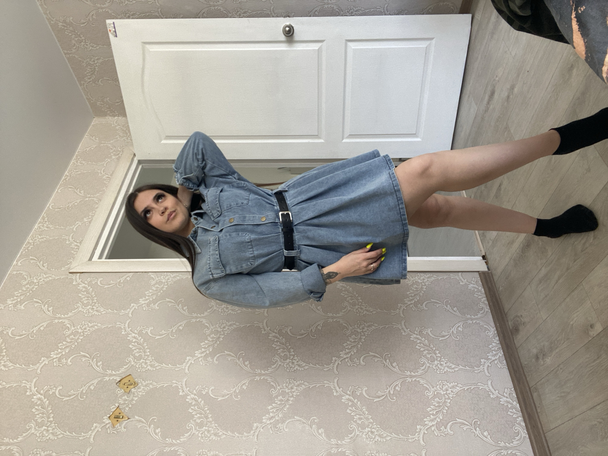 A woman is lying on the floor in a room, wearing a denim dress and black socks. She is facing up toward a closed white door. The walls are decorated with floral wallpaper. The floor is made of light-colored laminate. The photo was taken indoors. The city of Chaikovsky, Russia is not explicitly evident from the image.