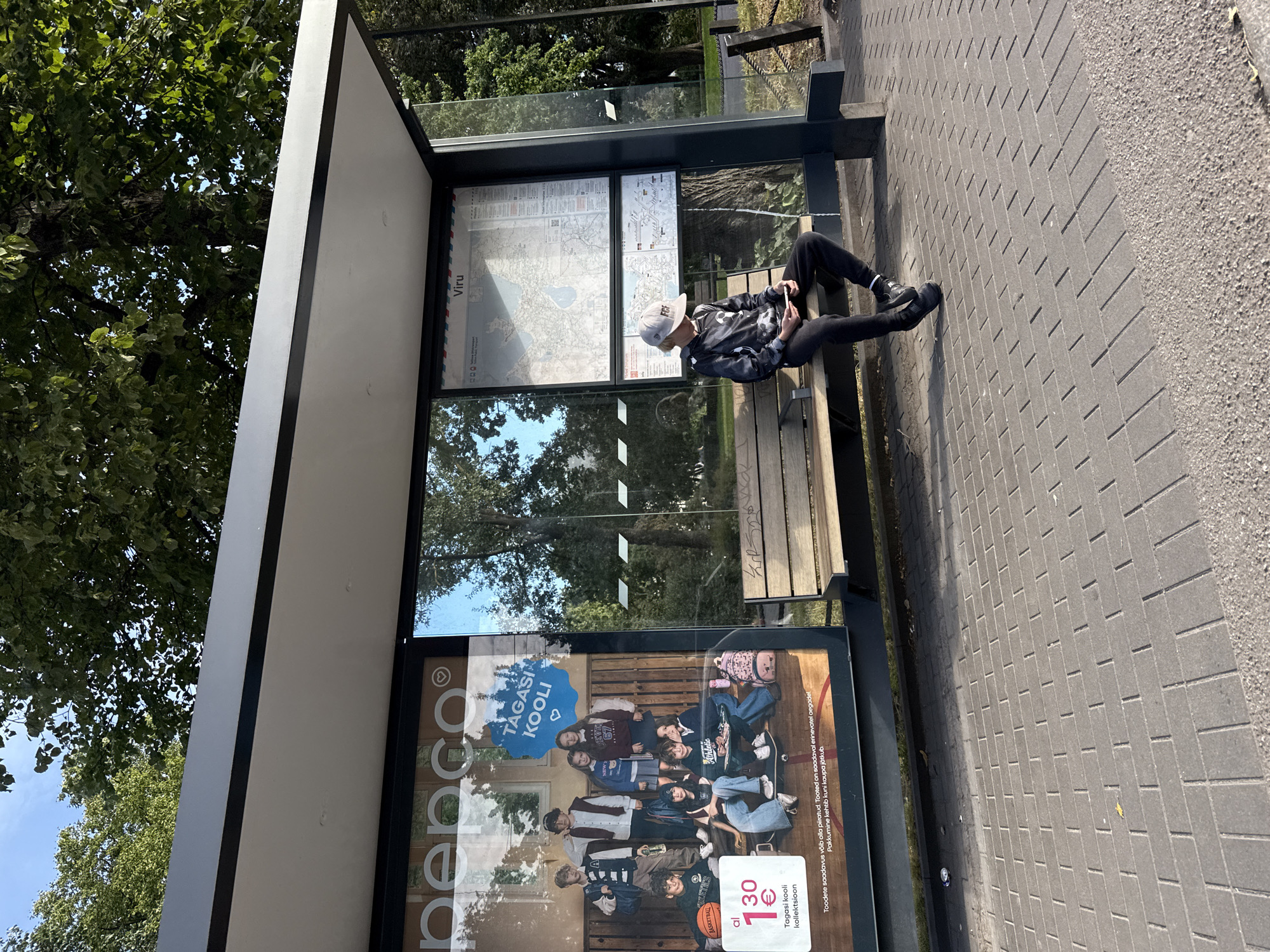 The image shows a bus stop with a young person sitting on the bench and looking at their phone. The bus stop has a large advertisement on the left side for "Pepco" and a map on the right side labeled "Viru". The young person is wearing a baseball cap, a dark hooded sweatshirt, black pants, and black sneakers. The bus stop is located on a paved sidewalk next to a street lined with trees. The sky is blue with some clouds.