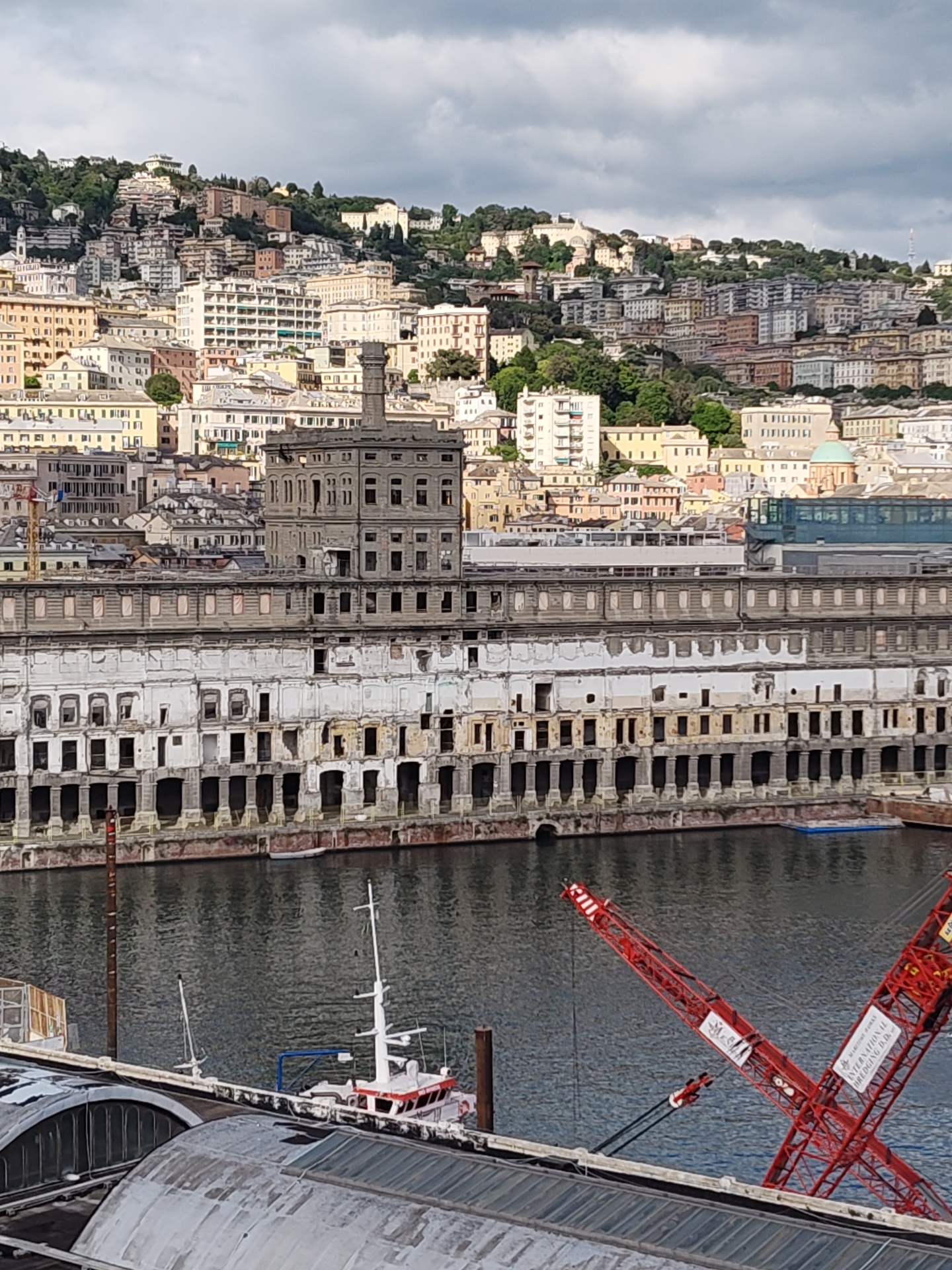 The image depicts a view of Genoa, Italy, focusing on a large, dilapidated building along the waterfront. The building has multiple levels, with many empty window openings, giving it a derelict appearance. A structure on top of the building resembles a tower or a tall chimney.
In the foreground, part of a metallic roof with a curved, industrial design is visible, along with a small white boat. A red crane is partially visible in the lower right of the image.
The background shows a hillside densely populated with buildings, indicative of the urban landscape of Genoa. The sky is overcast with cloudy conditions. The water in the foreground is calm.