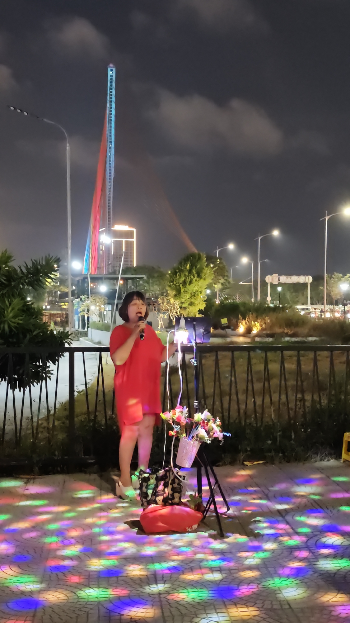 An outdoor night scene in Da Nang, Vietnam, shows a woman in a vibrant red dress singing into a microphone. She stands on a paved area brightly illuminated by a swirling pattern of multicolored disco lights in hues of red, green, blue, and yellow. To her right, a stand holds what appears to be a speaker or amplifier, adorned with a white pot overflowing with colorful artificial flowers. A red bag or cushion rests on the ground near the stand.
Behind the woman is a black metal fence, beyond which lie various trees and bushes, leading to a busy city road. The background is dominated by a towering, slender structure, characteristic of the main pylon of the Dragon Bridge, glowing with alternating blue and red light against the dark, cloudy night sky. Other city buildings with illuminated windows and numerous streetlights contribute to the urban glow. Distant vehicle lights suggest active traffic. The woman appears focused on her performance, her mouth open in song. No clearly legible text is visible on any signs or objects.