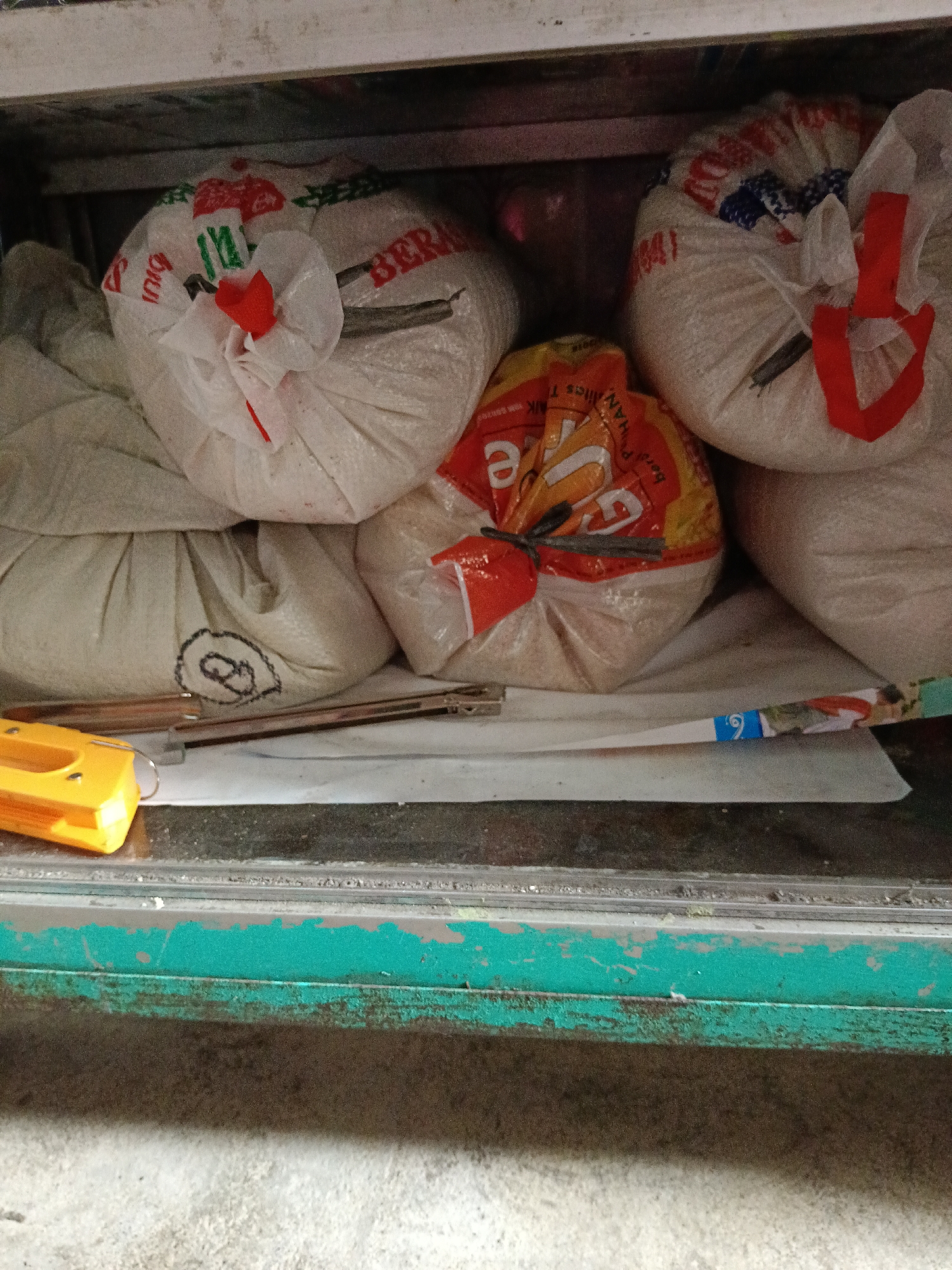 The image shows a close-up view of several large bags of rice stacked in what appears to be a storage area. The bags are made of white and patterned plastic, and some have red ties. One bag in the foreground has an orange and yellow label with some text visible, including what looks like "BERAS" (rice in Indonesian) and some Asian characters. To the left of the bags, a bright yellow stapler-like object is partially visible, resting on a white surface. The background is dark and indistinct, suggesting an indoor, possibly dimly lit space. The overall impression is of a stock of goods in a commercial or storage setting, likely in Aek Kanopan, Indonesia, given the context provided. The image is taken from a slightly elevated, downward angle. The lower part of the image shows a green, weathered metal surface.