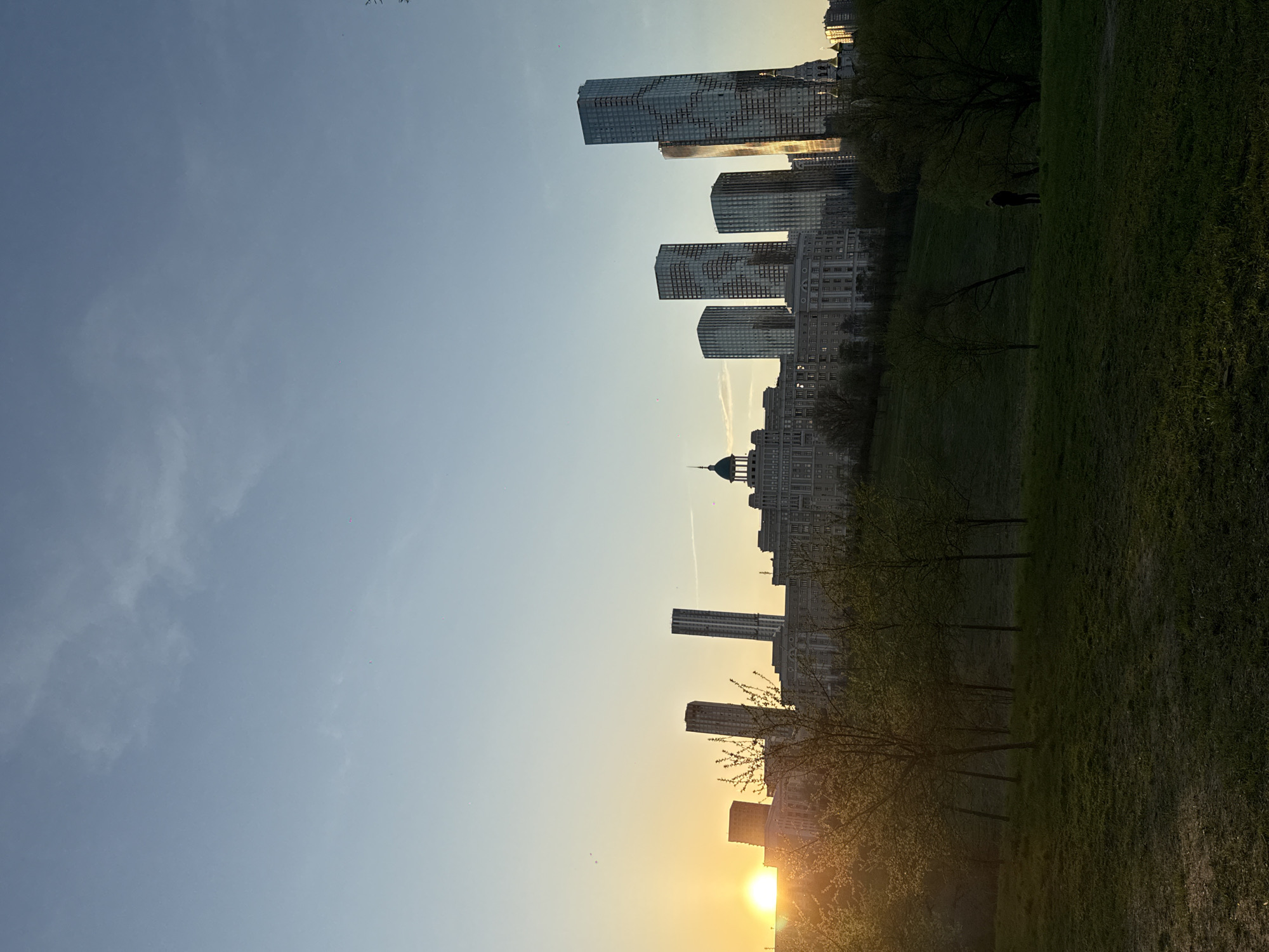 The image captures a cityscape at sunset, likely in Moscow, Russia, as suggested by the presence of the Moscow International Business Center skyscrapers. The scene is split between a clear sky transitioning from blue to golden hues at the horizon and a green grassy area in the foreground.
The Moscow City complex consists of several tall skyscrapers, including buildings with mirrored glass facades, suggesting commercial or residential use. There are also low-rise buildings with detailed architectural features near the skyscrapers, likely historical or residential in nature.
In the grassy area, there are trees with delicate branches and emerging leaves, indicating the early stages of spring. A dark figure is visible in the distance on the grass, possibly an individual walking or standing in the park.