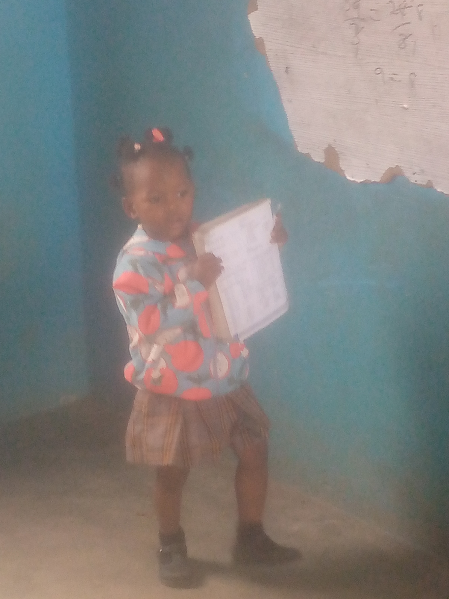 A young girl stands in a room with blue walls and a worn concrete floor. She is holding a small whiteboard that has what appears to be letters on it. She is wearing a patterned long-sleeved shirt and a plaid skirt, with dark-colored shoes. On the wall to the right of the girl, there is a damaged whiteboard with faded writing visible on it. The location is Unknown, Unknown.
