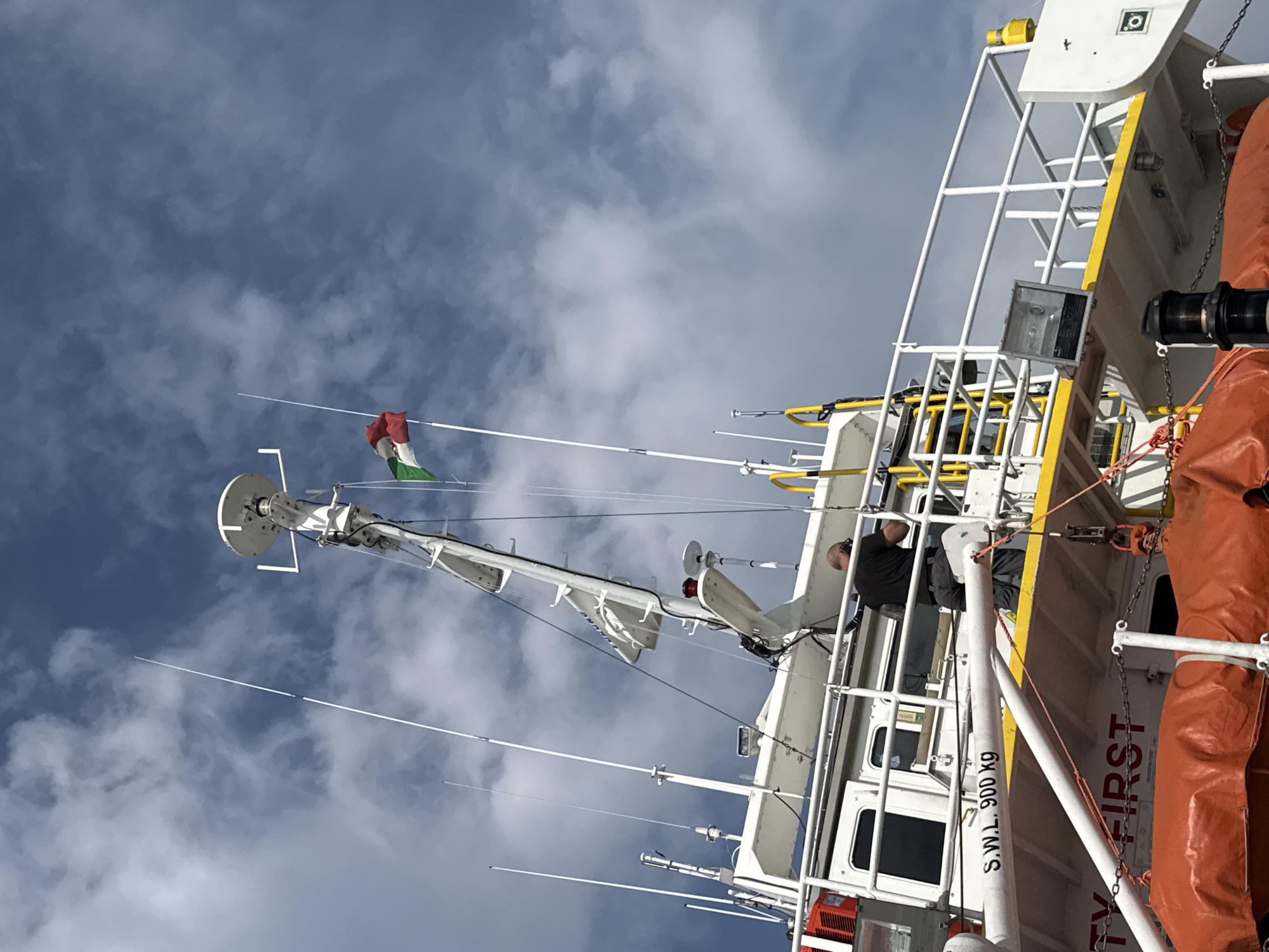 The image depicts the upper deck of a vessel docked in Marina di Ravenna, Italy. A person wearing a black t-shirt and dark pants is visible, standing on the deck and looking down at the ground. They appear to be wearing headphones. The vessel features a white superstructure with various antennas and communication equipment mounted on masts. A flag with green, white, and red horizontal stripes, likely the Italian flag, is flying from a mast. A white bar with black lettering reads "S.W.L. 900 KG." Below this, the words "TY FIRST" are partially visible in large red lettering. Several orange life rafts are secured on deck. The sky is blue with scattered clouds.
