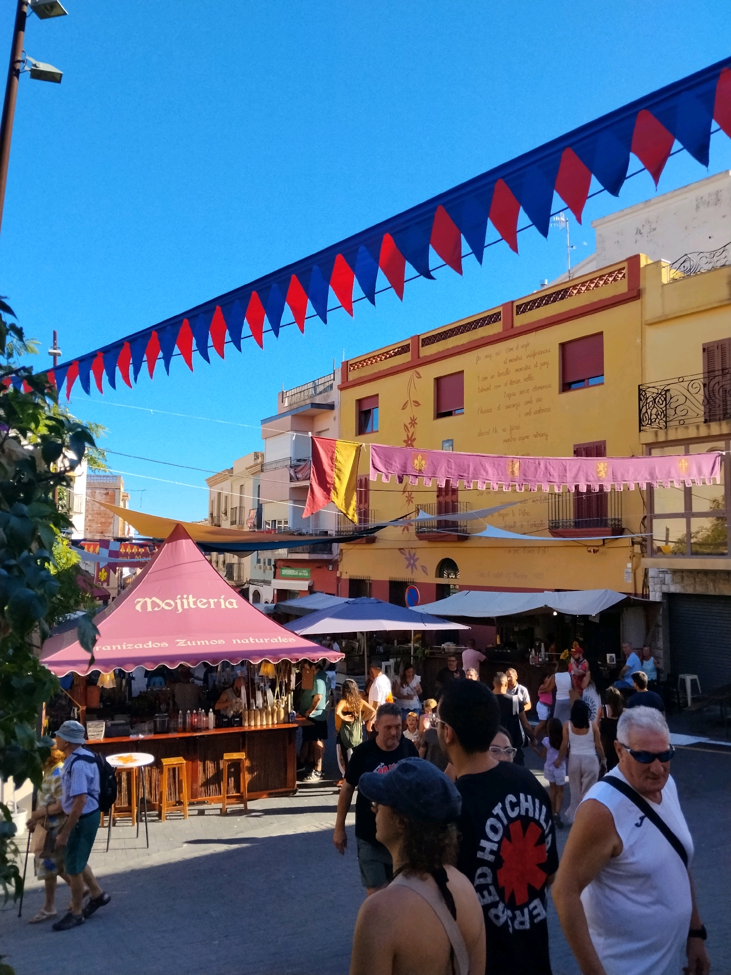 The image depicts a sunny street scene in Calafell, Spain, likely during a festival or market. Red and blue pennant banners are strung across the street, suspended from poles and buildings. Buildings on either side of the street are visible, with yellow and other pastel colored facades. Several market stalls are set up, including a prominent pink tent labeled "Mojitería" which offers "Organizados Zumos naturales" (Natural Juices). People are walking through the street, some browsing the stalls, while others are standing or sitting. In the foreground, a man wearing a white tank top and sunglasses walks away from the camera, while a couple walks towards the camera. The overall atmosphere appears lively and festive.