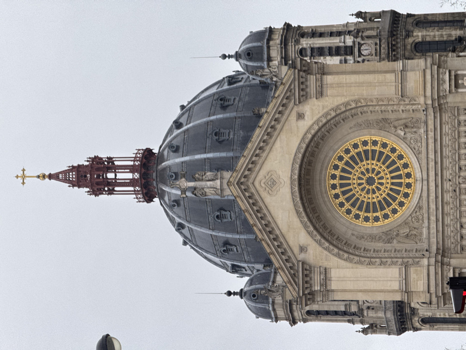 The image displays the upper facade and dome of the Église Saint-Augustin de Paris, located in the 8th arrondissement of Paris, France. The scene is captured under an overcast sky, presenting a uniform pale grey backdrop.
The central feature is a large, segmented dome, dark grey in color with lighter grey structural elements and small decorative architectural motifs resembling gabled lucarnes. Atop the dome is an ornate, deep red wrought-iron lantern and spire, culminating in a prominent golden Latin cross. Below the dome, on the main facade of the church, a large, circular golden rose window with intricate tracery is centrally positioned within a grand arched recess. Statues are visible carved into the stone beneath the rose window. The facade itself is constructed from light-colored stone, featuring decorative moldings, columns, and a pediment above the arch containing the rose window.
Flanking the central structure are two smaller towers, partially visible. The left tower features a small dark dome topped with a lightning rod. The right tower displays a visible clock face and additional architectural detailing, also topped with a smaller dark dome and lightning rod. A portion of a street lamp is visible on the far left edge of the frame, and a hint of dark tree foliage is present at the bottom right. No people or specific actions are discernible within the frame.