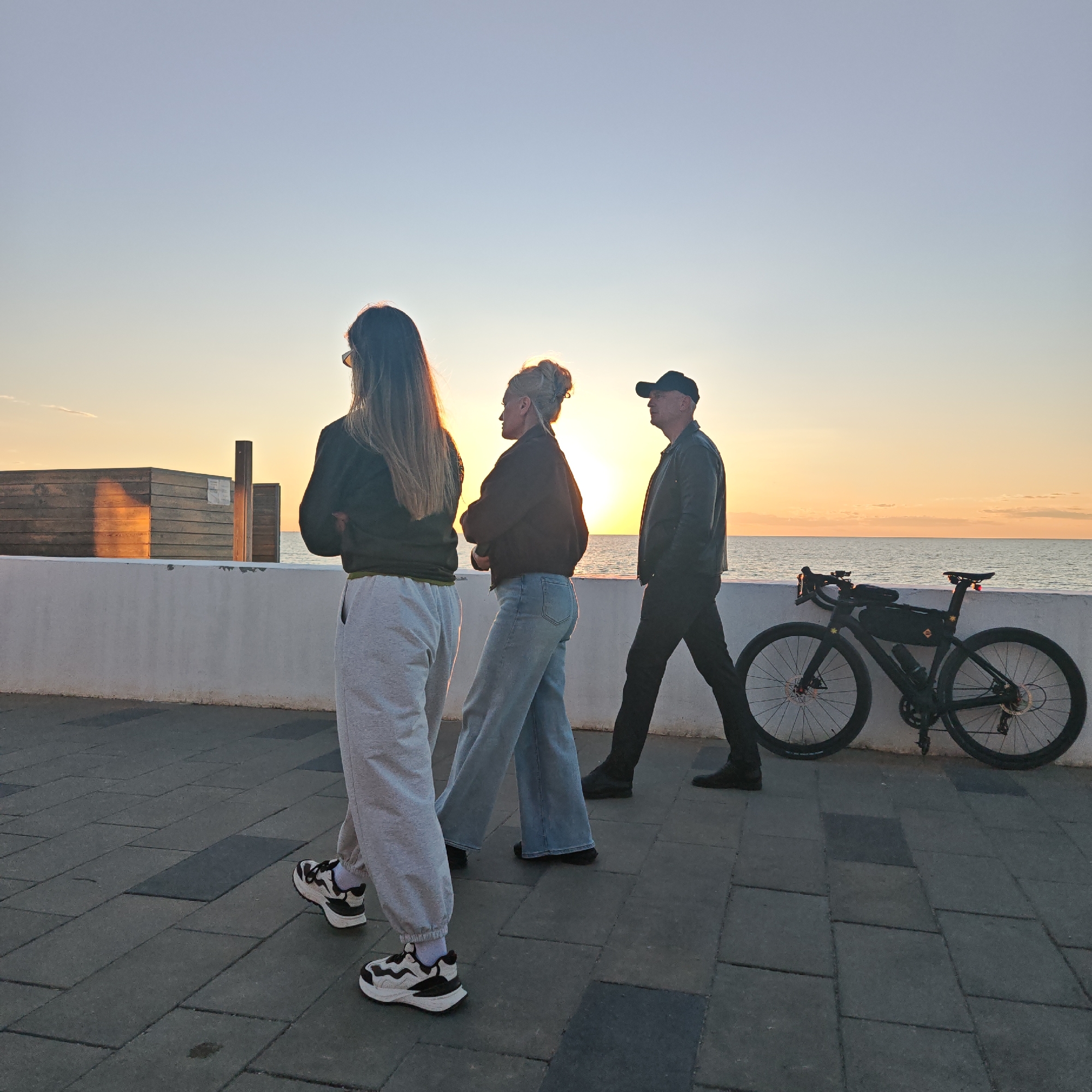 The image captures an outdoor scene at sunset, likely along a coastal promenade or boardwalk. Three individuals are visible, silhouetted or backlit by the setting sun over a body of water.
On the left, a woman with long dark hair, wearing sunglasses on her head, a dark jacket, light grey sweatpants, and white sneakers, stands with her back to the viewer, facing slightly right. Her arms appear crossed. In the middle, another woman with light hair styled in a bun, dressed in a dark jacket and light blue wide-leg jeans, is seen in profile, facing right. On the far right, a man wearing a dark baseball cap, dark jacket, dark trousers, and dark shoes walks towards the right, also seen in profile.
A black road bicycle with disc brakes, a frame bag, and a water bottle is parked or leaning against a white concrete wall that runs horizontally across the mid-ground, separating the paved path from the calm ocean or sea. The sun, a bright orange orb, is low on the horizon, casting a warm glow across the sky and reflecting on the water. In the background to the left, behind the white wall, there are some wooden structures, one of which shows a strong reflection of the golden sunlight. The ground is a paved surface with grey and darker rectangular tiles.
The time of day is clearly sunset, creating a dramatic and serene atmosphere. The weather appears clear with no significant clouds. No discernible text is visible on any of the subjects or objects in the image.