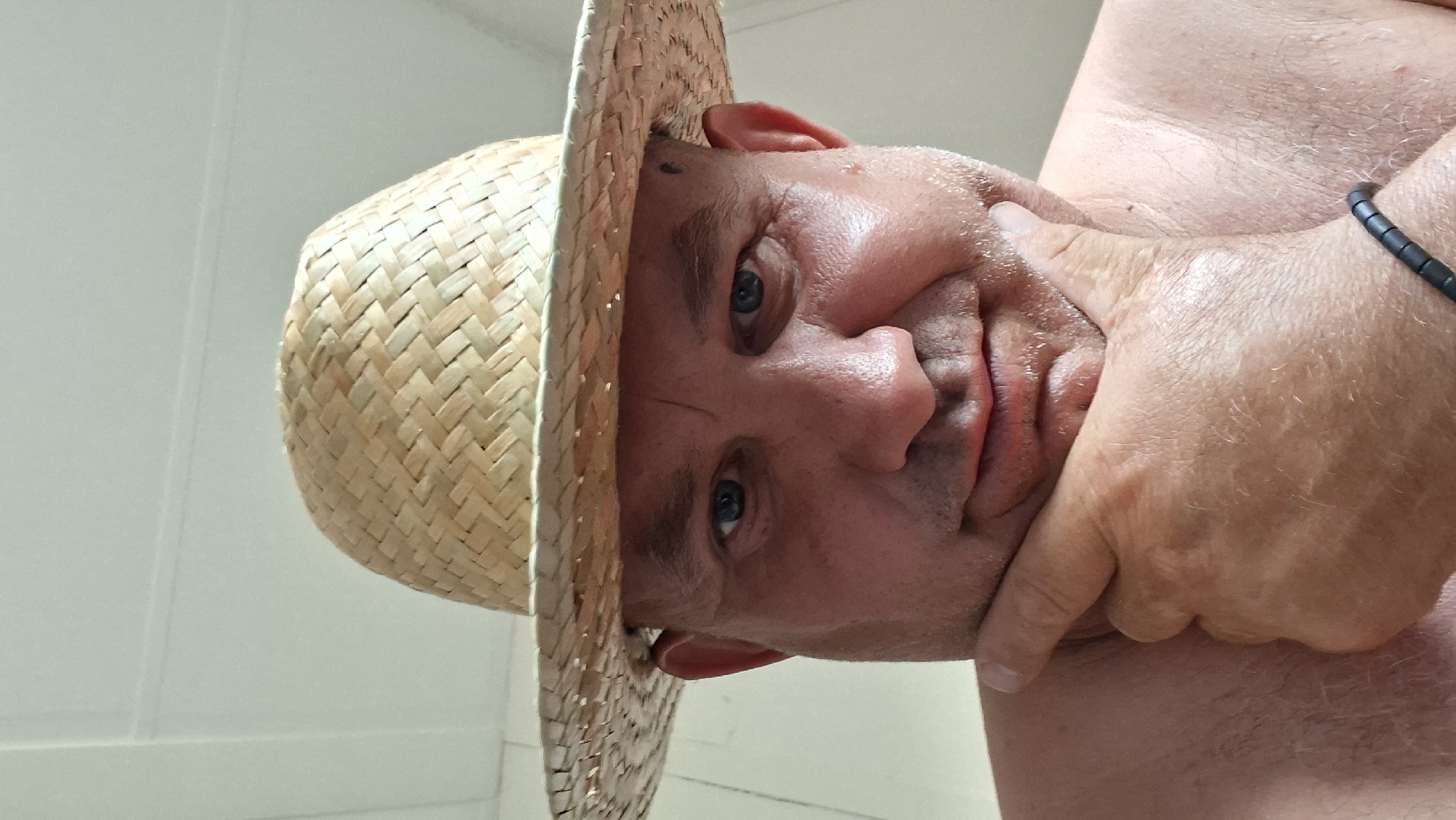 This is a close-up, front-facing selfie of a man wearing a straw hat. He has fair skin, blue eyes, and appears to be middle-aged with stubble on his chin and a mole on his right cheek. His right hand is resting on his chin, and he is looking directly at the camera with a slight smile. The background consists of a light-colored, vertically paneled wall. There are no indications of the city of Hîncești, Moldova, in the image.