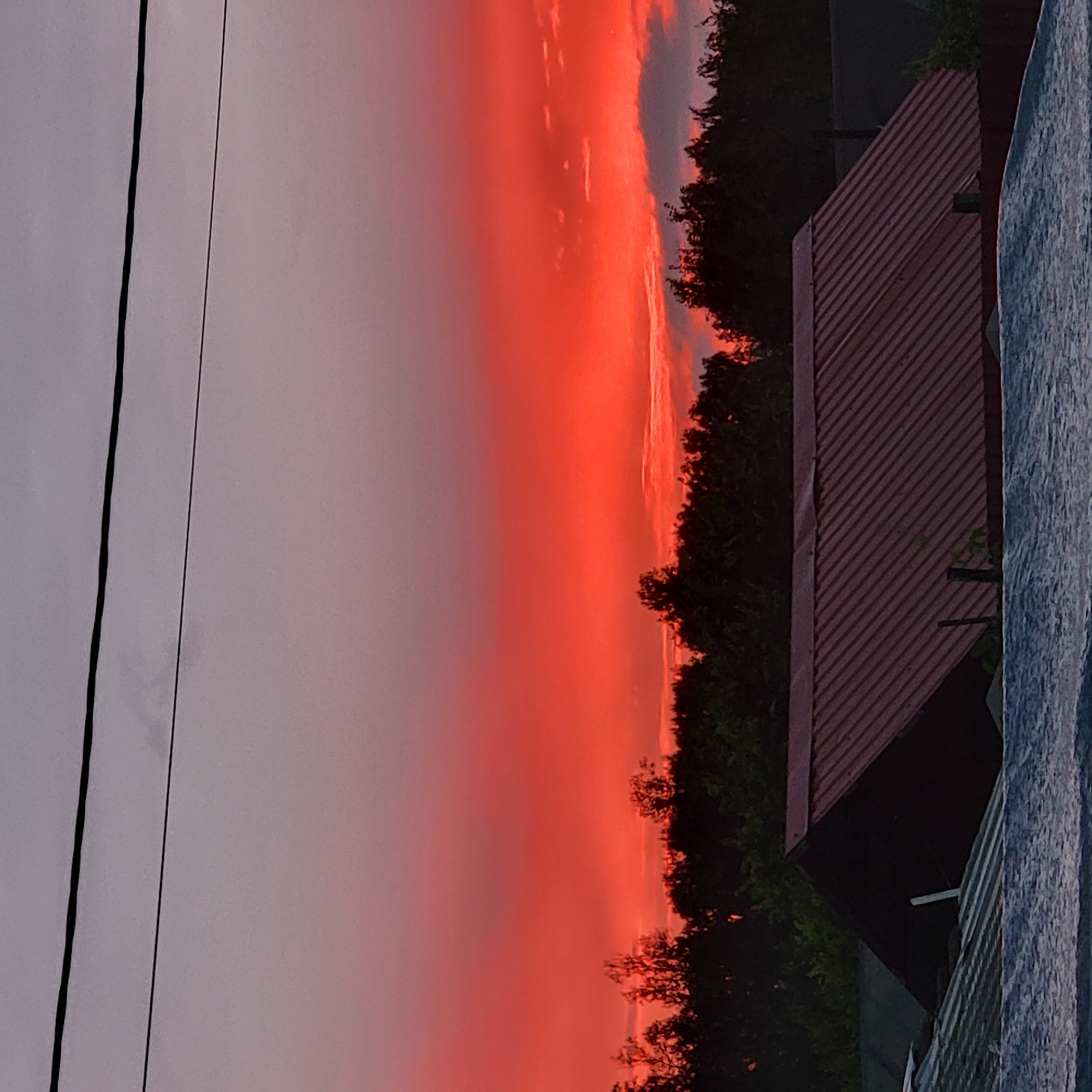 An outdoor, low-angle, medium shot captures a dramatic sunset over a rural area. The sky is ablaze with vibrant red and orange hues, transitioning to a lighter purple and grey at the top. Two dark power lines bisect the upper portion of the frame horizontally. Silhouetted against the colorful sky is a dense line of dark trees. In the foreground, the roofs of several buildings are visible. The most prominent is a red-metal, gabled roof with a series of parallel ridges. To its left, a darker, angular roof is partially visible. In the very bottom foreground, a rippled, silvery surface, possibly a body of water or a textured roof, dominates the lower part of the image. The scene appears to be in the city of Iglino, Russia, and is devoid of any human presence.