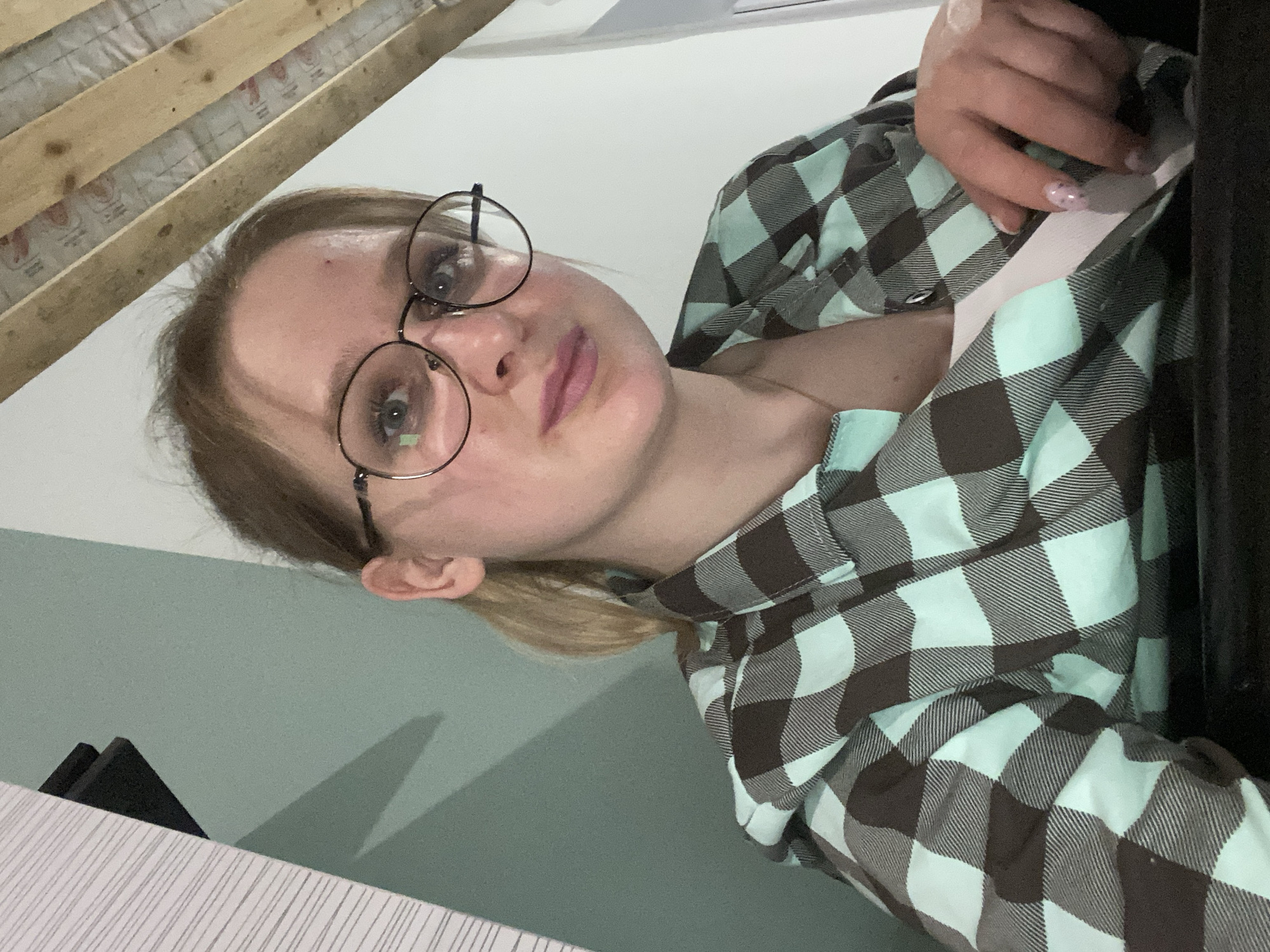 The image shows a woman with blonde hair pulled back in a ponytail, wearing round black glasses and a checkered flannel shirt in shades of brown and light blue. She is holding a white object with her right hand, which is slightly blurred. The background consists of a pale green wall and a wooden structure in the upper left corner. A black object sits on a white striped surface in the lower left of the frame. The image does not provide information about the location in the city of Unknown, Unknown.