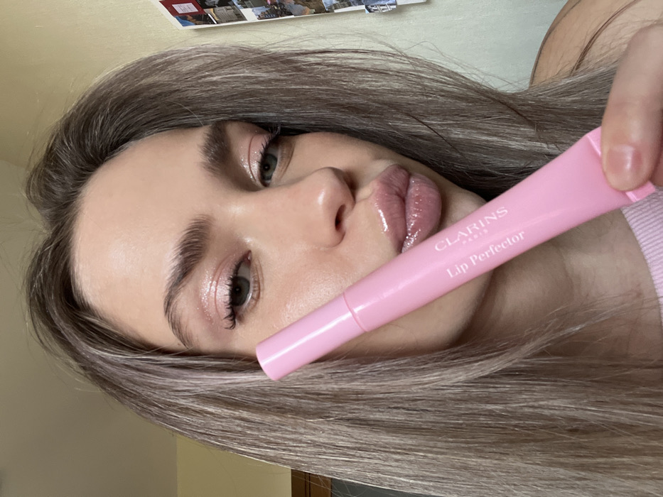 An individual, a woman with light brown hair and prominent eyebrows, is depicted in a close-up, chest-up shot. Her eyes are open and directed towards the camera, and her lips are slightly pursed in a pout, covered with a high-shine gloss. She wears light pinkish eyeshadow with mascara. Her right hand, partially visible in the lower right, holds a light pink tube of "CLARINS PARIS Lip Perfector," angled upwards from the lower right of the frame towards her face. The top of a light pink garment is visible around her neck. The background shows an indoor environment. To the left, a light-colored wooden door frame or wall section is visible. To the right, a plain light-colored wall features several small rectangular items, appearing to be photos or postcards, attached in an informal collage. The scene is an indoor setting in Saint Petersburg, Russia.