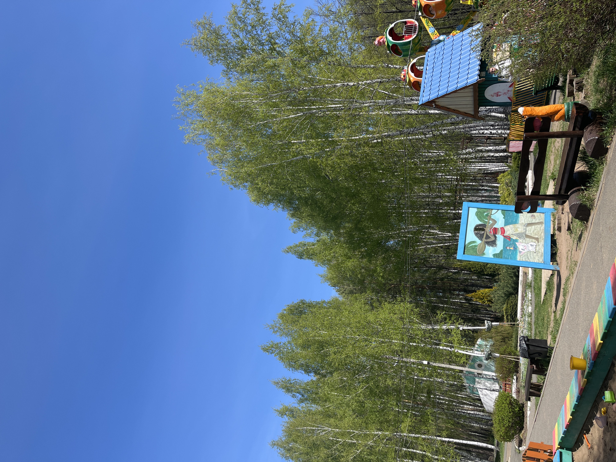 The scene presents a recreational area in Chaikovsky, Russia. A dense row of birch trees with light green foliage lines the center, contrasting against a clear, bright blue sky that dominates the left portion of the image. Along the right side, there's a glimpse of an amusement park ride with brightly colored cabins. Closer to the foreground on the right, a display includes a small, house-like structure with a blue roof, a bench, and a framed picture depicting a scene with cartoon-like characters on a beach. A colorful fence marks the edge of what appears to be a sandbox. The overall setting suggests a park or children's playground on a sunny day.