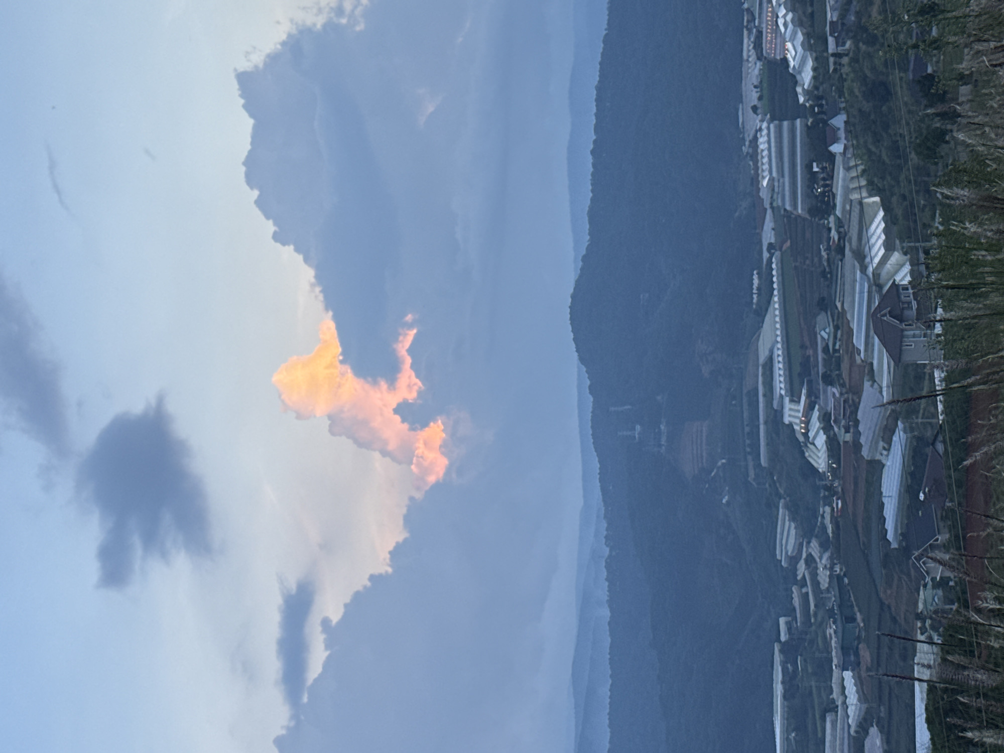 This is a vertical, eye-level shot of the Đa Lộc, Vietnam area at dusk. The sky is a soft blue with scattered clouds, some of which are illuminated by the setting sun with a vibrant orange and pink hue. In the middle ground, rolling hills covered in dark green vegetation recede into the distance. The foreground is dominated by a village nestled in the valley, characterized by numerous greenhouses with white, translucent roofs and a few houses with dark roofs. The overall atmosphere is serene and peaceful, with the fading light casting a gentle glow over the landscape.
