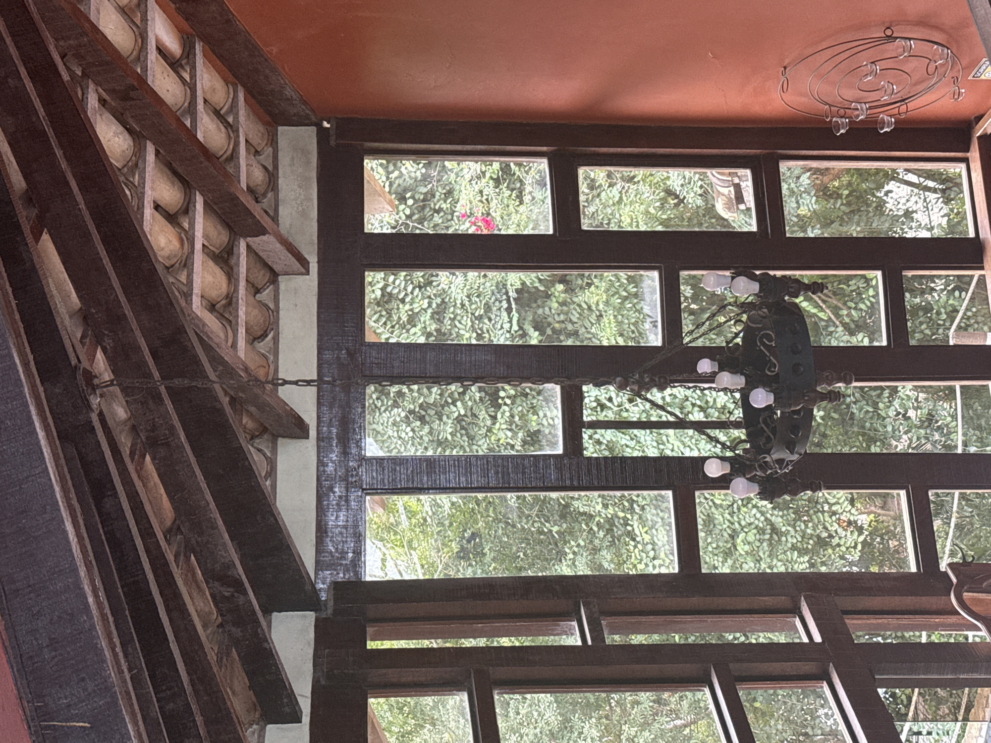 The image shows an interior view of a room with a rustic aesthetic, likely a house or restaurant, with large windows offering a view of lush green foliage. The ceiling features exposed dark wooden beams supporting clay roof tiles. A dark, ornate chandelier with several light bulbs hangs in the center of the frame. The walls are a warm, terracotta orange. To the right, a circular metal wall decoration with candle holders is visible. The overall impression is one of a relaxed, nature-integrated living space. The provided information about the city of São Pedro Da Aldeia, Brazil, suggests this scene is located there, but there are no visual cues in the image to confirm this specific location. There are no people or actions depicted in the image.
