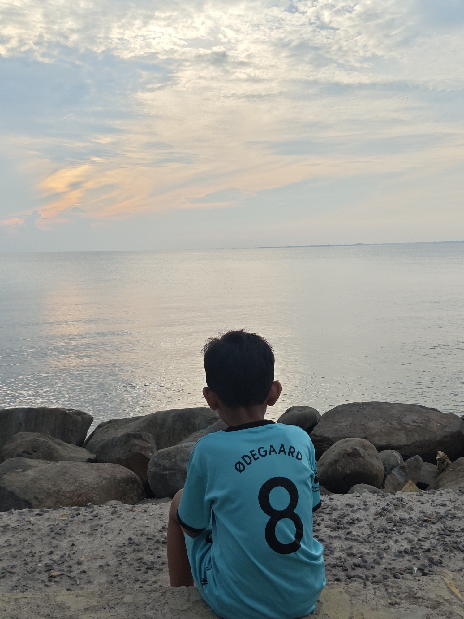 A young boy, viewed from the back, sits on a rocky shore, facing a calm sea under a cloudy sky at dusk. He wears a light blue t-shirt with black lettering that reads "ØDEGAARD" and the number "8". The sea is vast and reflects the muted colors of the sky, which has streaks of orange and yellow from the setting sun. Large, grey rocks are scattered on the shore in front of and around the boy, interspersed with sand and small pebbles. In the distance, a faint line of land is visible on the horizon. This scene is likely located at the beach of Ujong Blang in Lhokseumawe, Indonesia, a popular spot for its serene views.