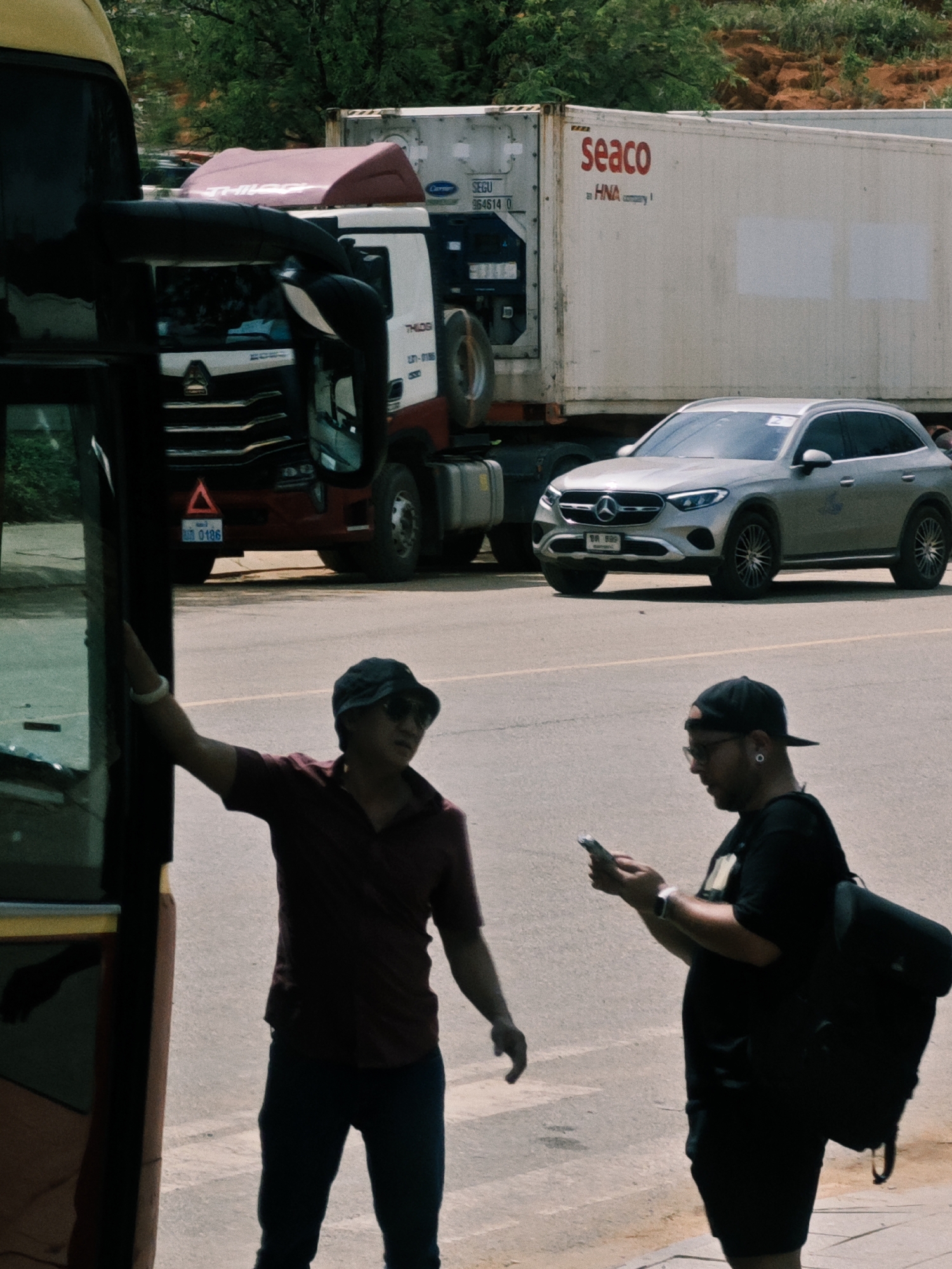 In Bờ Y, Vietnam, this image captures a scene near a roadside with several vehicles. A large yellow and red truck, possibly a bus or transport vehicle, is parked on the left, partially obscuring a man in a maroon shirt and dark pants. He is wearing a dark baseball cap and sunglasses and is leaning against the vehicle, holding a white bracelet on his left wrist. To his right, another man in a black t-shirt, black shorts, and a black baseball cap is looking at his mobile phone. He has earrings and a watch on his left wrist and is carrying a black backpack slung over his right shoulder. In the background, a large white shipping container with "seaco" branding is mounted on a truck. Further behind it, a silver Mercedes-Benz SUV is parked on the road. The ground is paved, and a patch of reddish-brown earth with green vegetation is visible in the upper background. The lighting suggests it is daytime.
