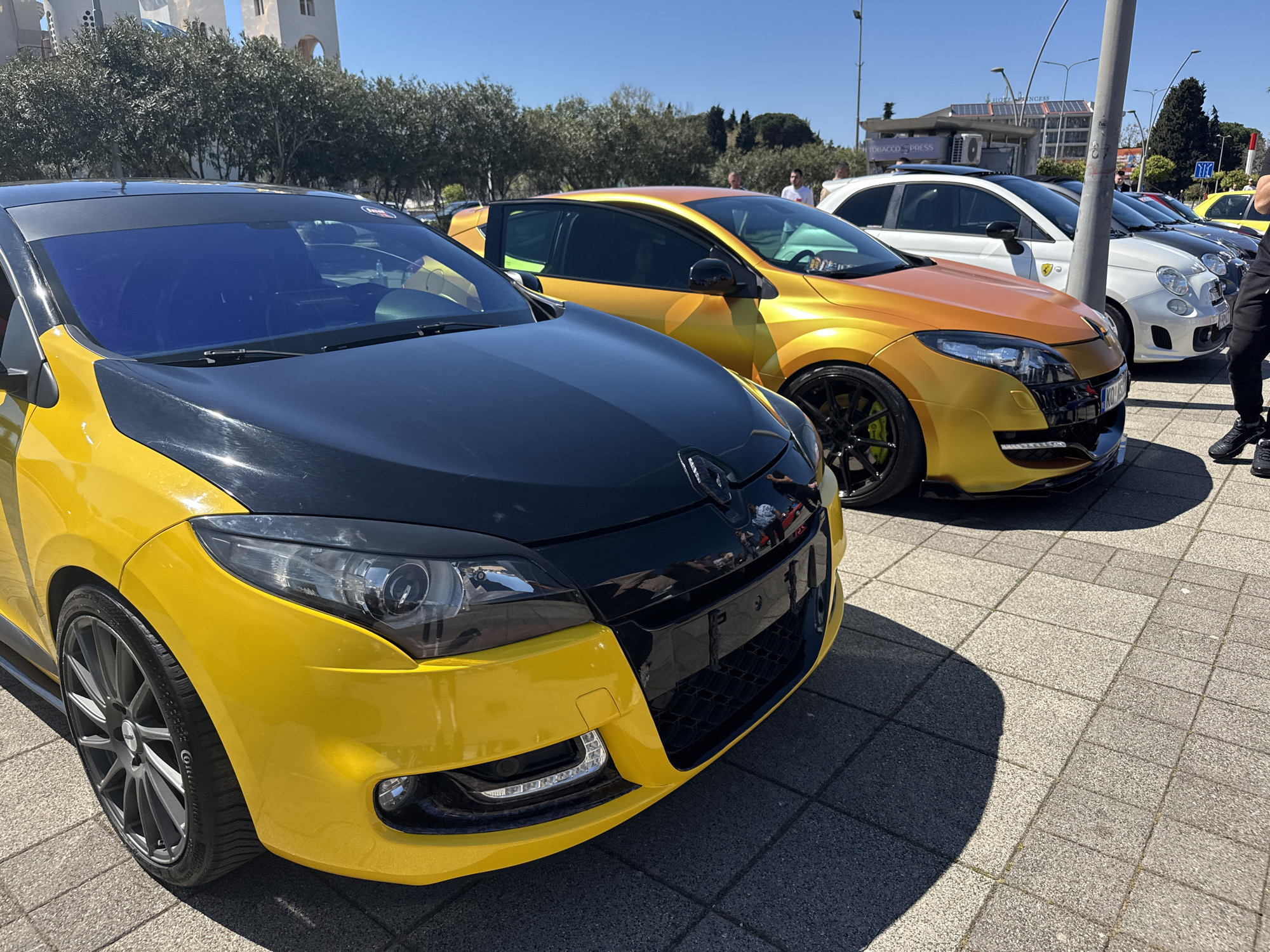 The image shows a row of cars parked on a paved surface in an urban setting. The foreground features a yellow and black Renault Megane, followed by an orange and gold Renault Megane. Behind them are several white Fiat 500s. A person is standing near the end of the row of cars on the right. Buildings and trees are visible in the background under a clear blue sky. A pole and street signs are also visible. There is no information available about the specific location in a city, and "Unknown, Unknown" does not provide valid geographic details.