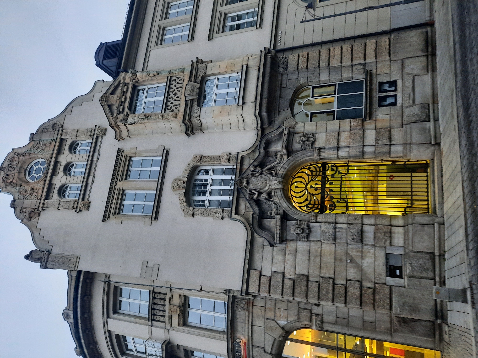 The image shows a detailed view of the exterior of the Sparkasse building in Rosenheim, Germany. The building features ornate architectural details with stone facades and decorative elements above windows and doorways. The main entrance is a large, arched gateway with intricate metalwork. The lower portion of the building is constructed of large, rough-hewn stone blocks, while the upper stories are rendered in a lighter color. Several windows of varying sizes and shapes are visible. The lighting suggests it is either early morning or late evening, with artificial lights visible inside the building. The street in front of the building is paved with cobblestones. There are no people visible in the image.