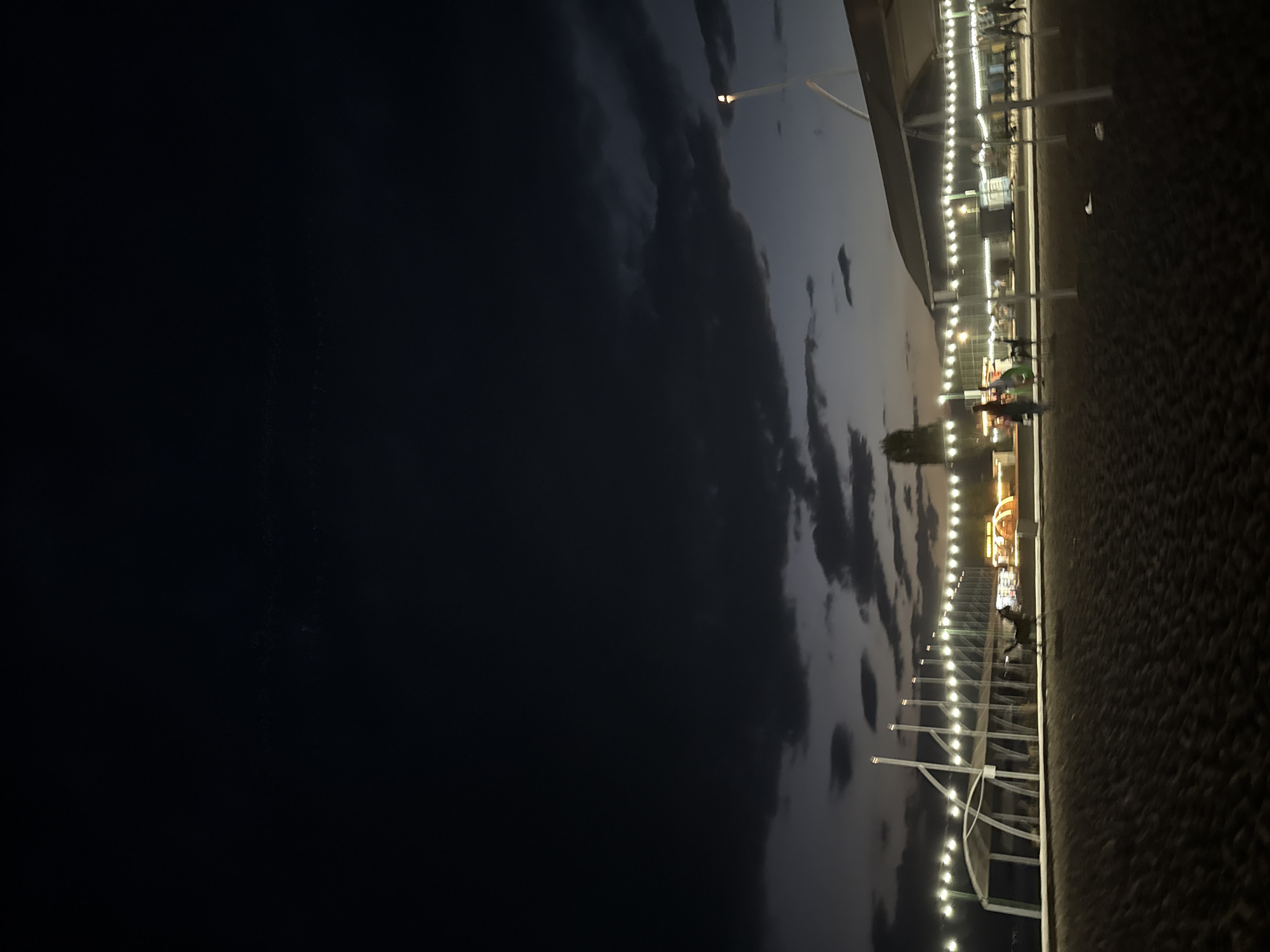 The image captures a dusk scene at a beach or waterfront area in Sary-Oy, Kyrgyzstan. A sandy or pebbly ground occupies the foreground. In the mid-ground, a row of white vertical poles supports a string of lights, creating an illuminated pathway or decorative border. Behind this, a covered structure with a white roof is visible, supported by similar poles. Further back, amusement park-like structures with colorful lights, including a Ferris wheel, suggest a recreational area. Several people are present, some walking along the illuminated path and others near the amusement structures. A horse is also visible in motion on the sandy ground. The sky above is dark with scattered clouds, and a few faint stars are discernible in the upper portion of the image, indicating the transition from twilight to night.