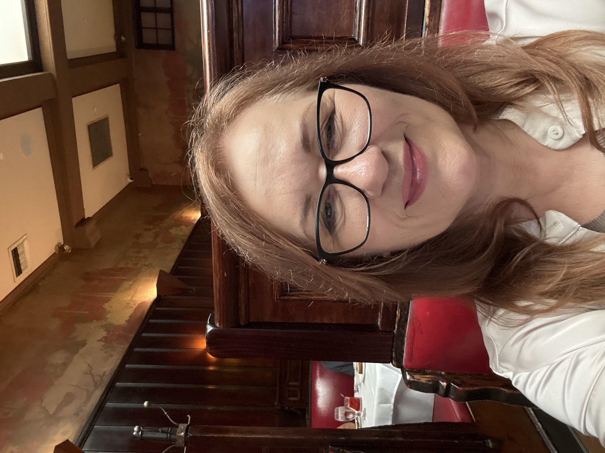 A woman with auburn hair and glasses is taking a selfie in a restaurant booth. The booth has red upholstery and dark wood paneling. In the background, a table is set with a white tablecloth, glasses of red liquid, and a plate. The restaurant has a warm, dimly lit ambiance, with wood accents on the walls and ceiling.