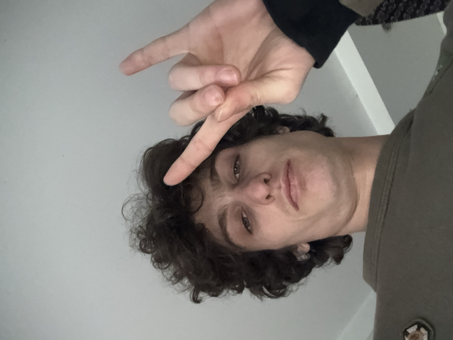 The image displays a headshot of a young individual, likely a male, rotated roughly 90 degrees clockwise, making the subject appear on their side with their head to the left and body extending to the right.
The person has dark, curly hair that appears to be medium in length, framing their face. Their skin tone is fair. They have distinct eyebrows, dark eyes that are looking directly towards the viewer, and a relatively straight nose. Their lips are closed, forming a slightly serious or neutral expression.
A hand is prominently featured in the upper left portion of the image (as viewed in its rotated state), making a "peace" or "V" sign. The index and middle fingers are extended upwards, while the ring finger and pinky are curled down, and the thumb is also tucked in. This hand belongs to the subject and is positioned above their head, with the extended fingers pointing somewhat towards the viewer. The wrist and part of an arm are visible, covered by a dark, possibly black, long-sleeved garment.
The subject is wearing a muted olive green or brown crew-neck top, visible on the right side of the frame. There's a small, textured emblem or patch of a lighter color (possibly gold or cream) with a brown outline visible on the chest area of the shirt.
The background is predominantly a plain, light grey or off-white surface, suggesting a wall or ceiling. There are no other discernible objects or details in the background, except for a very small, dark, pixel-like specks on the light background in some cropped versions, which are likely dust or minor imperfections.
The lighting appears even, suggesting indoor conditions, possibly with ambient light rather than direct harsh lighting. The overall mood is casual, with the hand gesture being a common pose for selfies or informal portraits. No text is visible in the image.