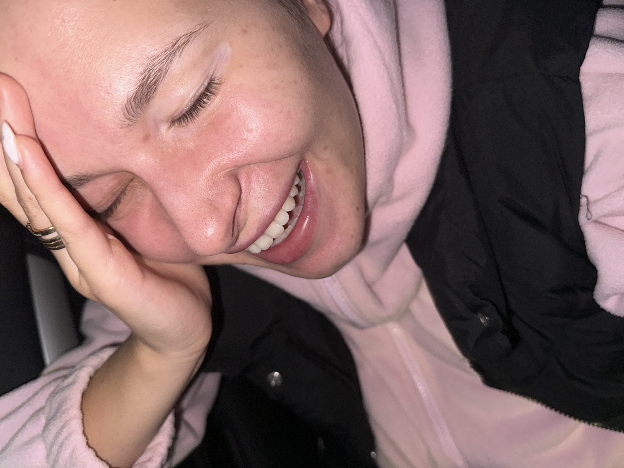A close-up, angled shot shows a fair-skinned individual with closed eyes, a bright smile revealing upper teeth, and their left hand partially covering their cheek. The person wears multiple rings on their fingers and has white nail polish on their long nails. A light pink hoodie is visible, layered beneath a black jacket. The environment appears dimly lit. There is no information about Filat'evo, Russia in the picture.