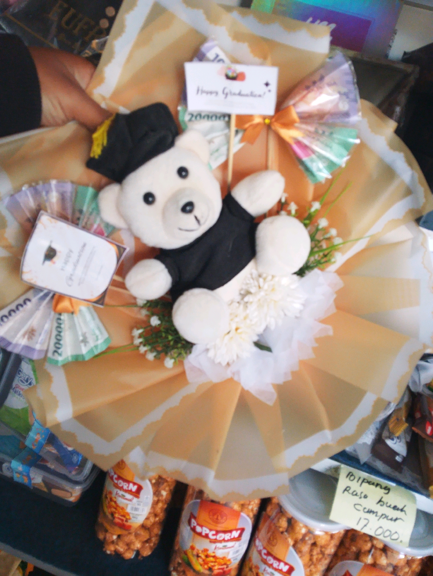 A graduation gift bouquet is displayed. The bouquet is made of a white stuffed teddy bear wearing a black graduation cap and gown. It is surrounded by fake white flowers and green foliage. The bouquet is wrapped in translucent orange paper and decorated with several folded Indonesian rupiah banknotes. A small white card with "Happy Graduation!" written on it is attached to the bouquet. Behind the bouquet, there are several bags of popcorn and other packaged snacks, along with a handwritten price tag that reads "17.000". The background suggests an indoor setting, possibly a stall or shop, with various items for sale. The overall scene indicates a commercial display of graduation gifts in Bangil, Indonesia.