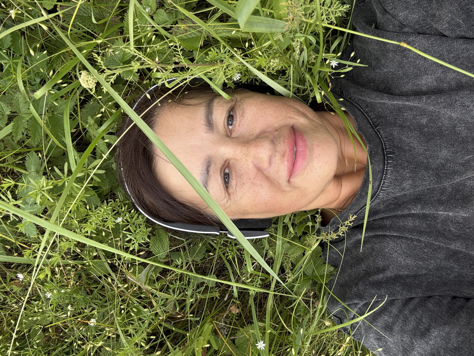 A woman with dark hair is lying on her back in the grass, wearing black headphones and a dark grey t-shirt. She is looking up at the camera, and her face is partially obscured by tall green grass. Her skin has freckles. The environment is natural, with various types of grass and small white flowers. The scene appears to be outdoors in a grassy area, possibly in the city of Kalinino, Russia, given the provided context.