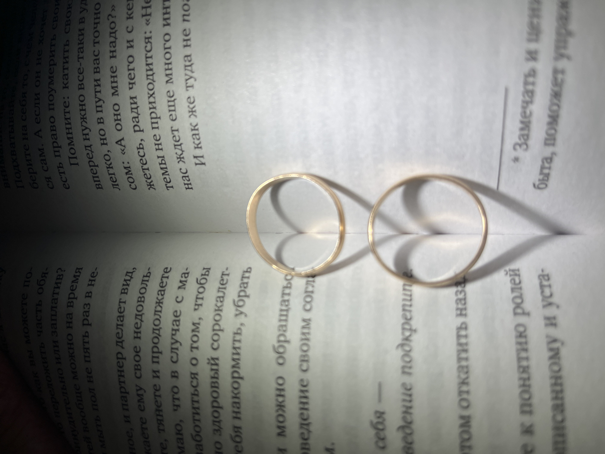 Here's a detailed description of the image:
**Overall Impression:** The image captures a close-up of two gold wedding bands resting open on the pages of a book written in Russian.
**Details:**
* **Objects:**
* Two gold wedding bands, arranged side-by-side.
* An open book with Russian text, providing a backdrop to the rings.
* **Text and Writing:**
* The Russian text is visible on both pages of the book. The text appears to be from a self-help or relationship advice book.
* **Environment/Lighting:**
* The setting is indoor, with the primary light source coming from above, casting shadows of the rings onto the book pages.
* **Color and Tone:**
* Warm tones, primarily from the gold rings and soft lighting.
* The pages of the book are a stark white, providing a contrast.
**Interpretation:**
* The image conveys themes of marriage, commitment, and perhaps relationships.
* The text in the background may be relevant to the symbolic meaning of the rings.
**Location (Chaikovsky, Russia):** There is nothing in the image that would explicitly confirm this photograph was taken in Chaikovsky, Russia. The presence of Russian text suggests the location is in Russia or a Russian-speaking region, but Chaikovsky cannot be determined with certainty based solely on the image.