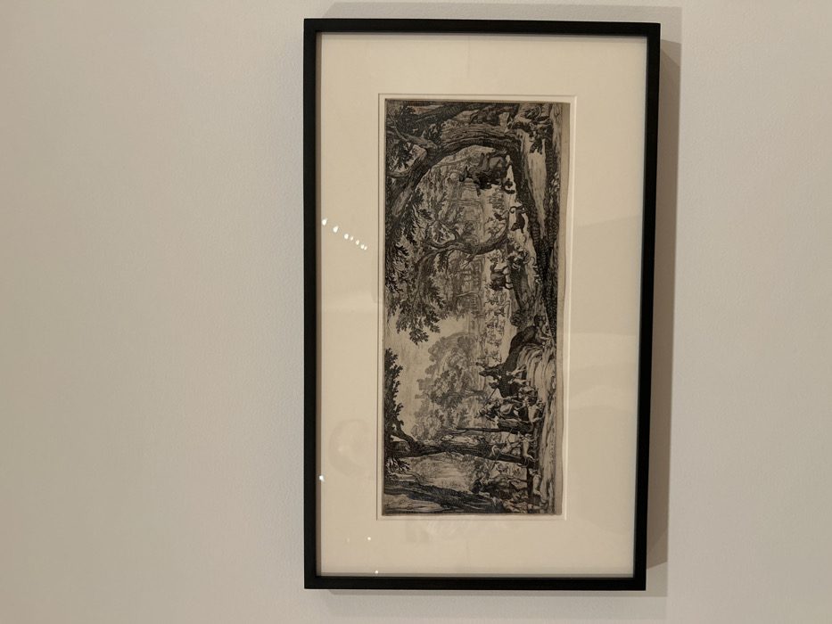 A horizontal, framed black-and-white engraving is mounted centrally on a plain, off-white wall. The artwork is encased within a sleek black rectangular frame, enclosing a wide, light-cream matboard. Minor specular reflections are visible on the glass covering the print.
The engraving depicts a dynamic, large-scale hunting scene set within a densely forested landscape with varied terrain. Numerous human figures, dressed in historical period attire, are actively participating in the hunt. Several individuals are mounted on horseback, while others are on foot, armed with spears and long-barreled firearms. A significant pack of hunting dogs (hounds) is prominently featured, engaged in the pursuit and attack of various animals. In the foreground, a violent struggle unfolds in a body of water or swampy area, where multiple crocodiles or alligators are being attacked by several dogs. Further into the midground, additional hunted animals, including deer and possibly wild boar, are visible amidst the commotion. Mature, heavily foliated trees frame both the left and right sides of the composition and extend into the background. In the far distance, a fortified structure, resembling a castle or walled town, is situated atop a prominent hill. The scene conveys intense action and widespread interaction between hunters, horses, dogs, and their prey.
This artwork is located within the Los Angeles County Museum of Art (LACMA) in Los Angeles, California, United States of America.