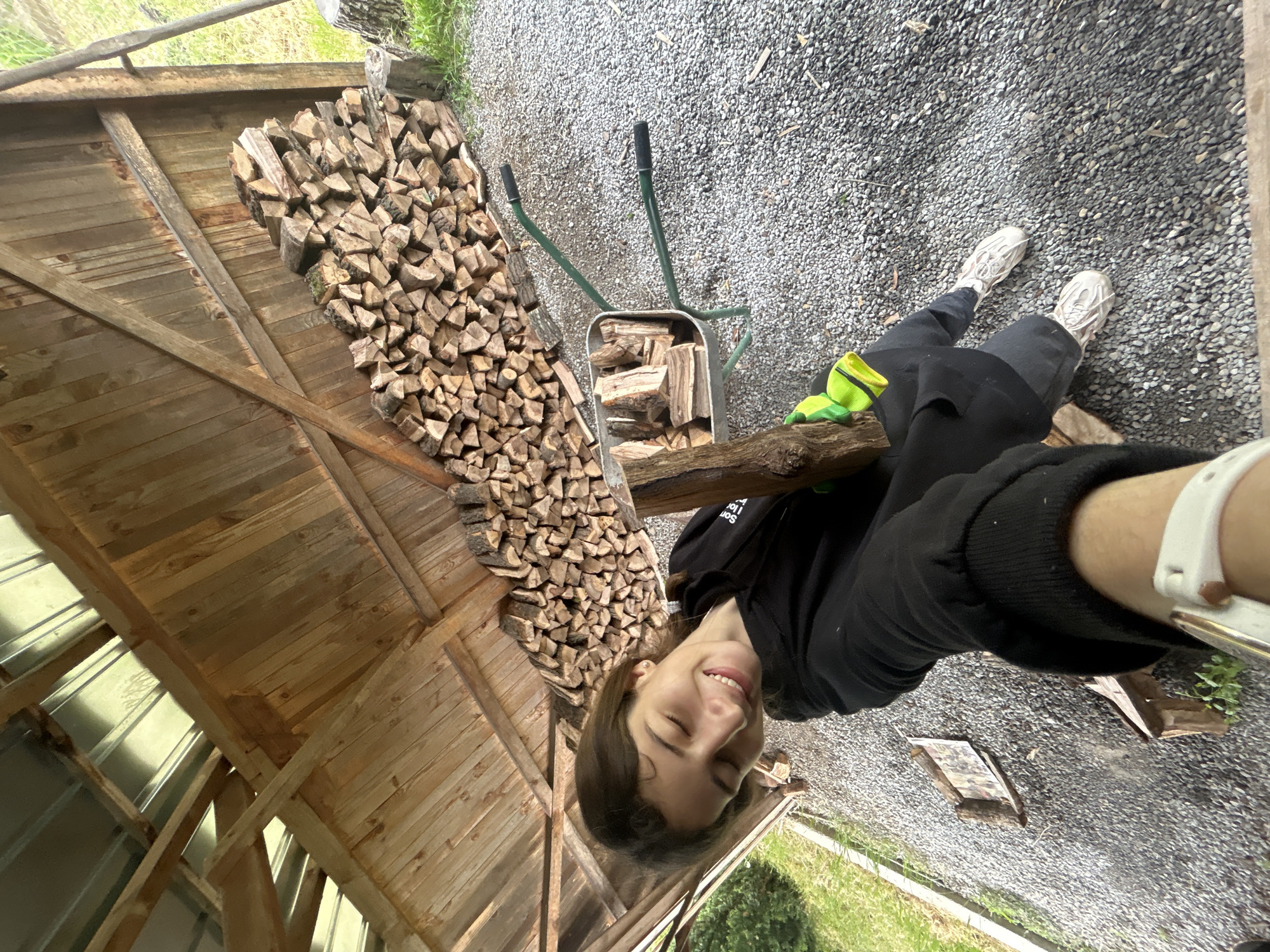 An outdoor scene in Senovo, Bulgaria, shows a smiling person in a black hooded sweatshirt, black pants, and white sneakers standing on gravel. A filled wheelbarrow sits nearby, and a large stack of chopped firewood is visible under a wooden structure. The environment includes gravel ground, grass, and a wooden building.