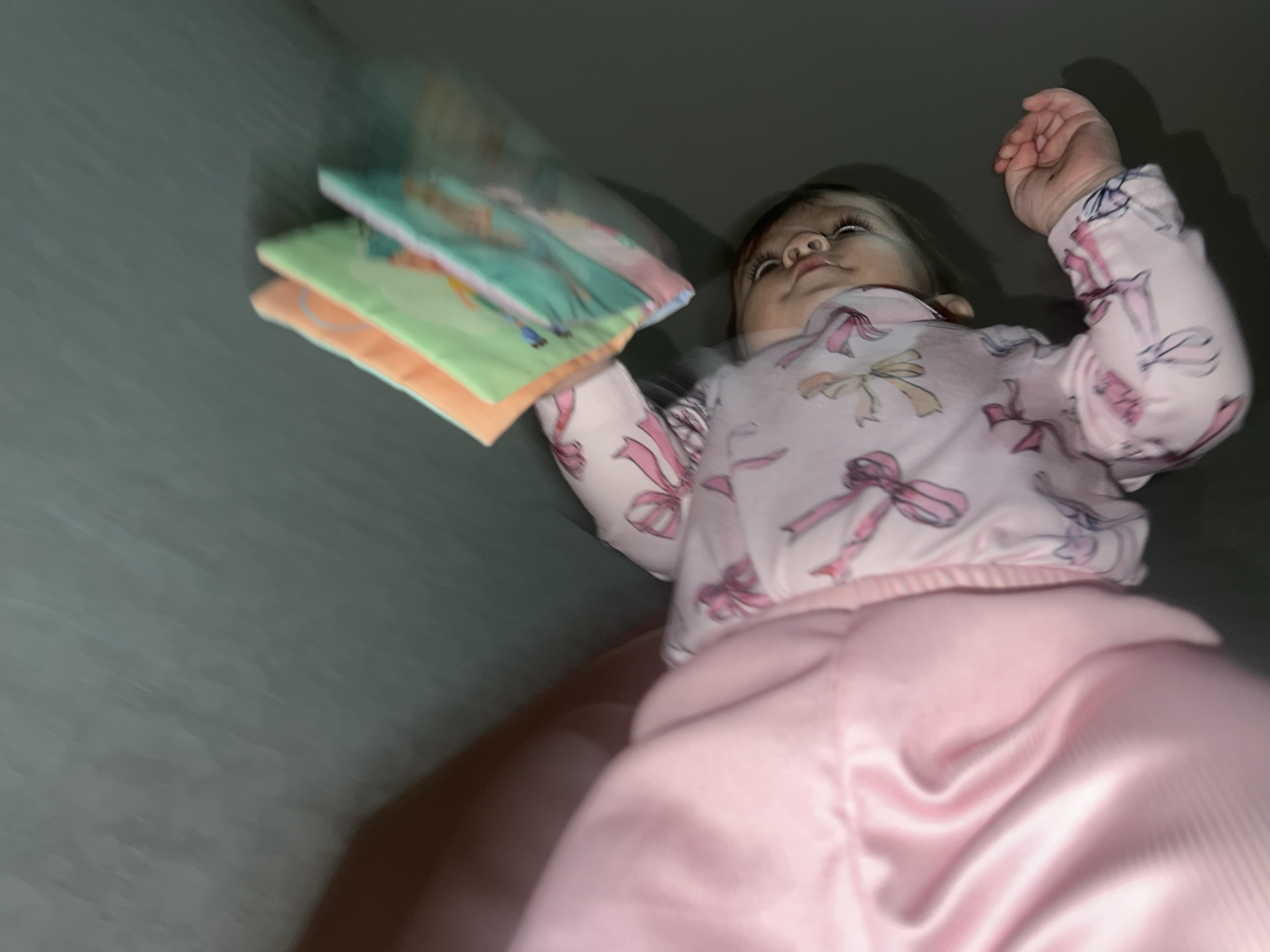 A low-angle shot of a baby wearing pink pajamas with a bow pattern. The baby is holding a stack of three colorful soft books. The setting appears to be indoors, with a gray wall in the background. The location details of Elena, Bulgaria, are not discernible from the visual content.