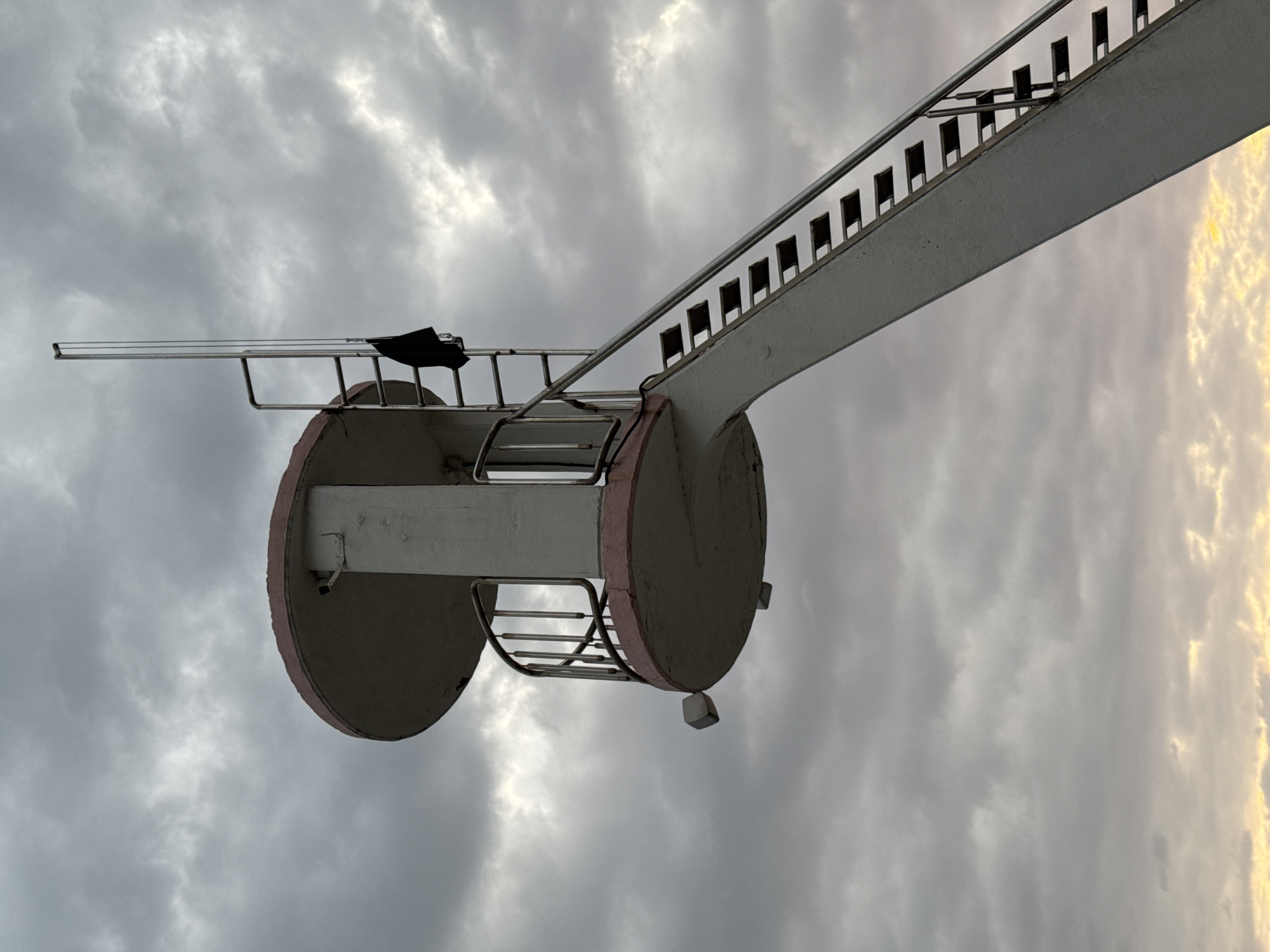 A concrete lifeguard tower with a pink trim stands against a cloudy sky in the Vardane Microdistrict, Russia. The tower has a circular platform at the top with a railing and a small enclosed area. A metal ladder extends from the platform upwards, with a black piece of fabric draped over it. A long, slanted staircase with metal railings leads up to the platform. The bottom of the image shows a sliver of yellow light on the horizon, suggesting either sunrise or sunset. The scene is devoid of people and activity.