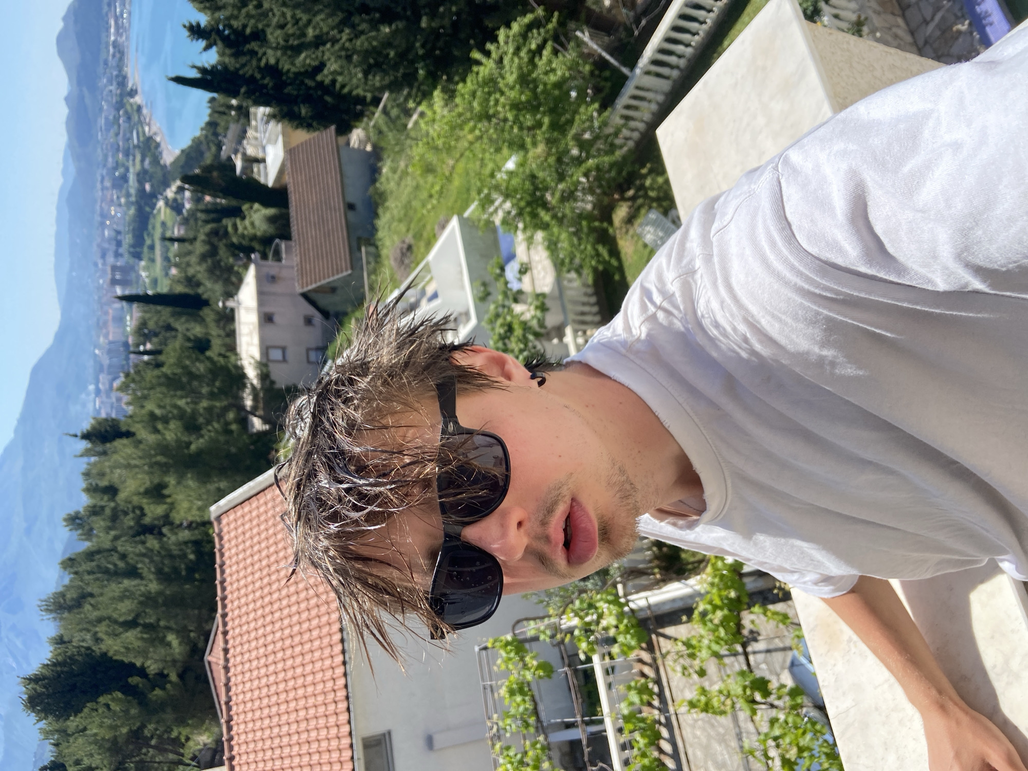 The image displays a selfie taken by a young man, rotated 90 degrees clockwise, placing the horizon vertically. The central subject is a young male, likely in his late teens or twenties, seen from the chest up. He wears a plain white t-shirt and dark sunglasses, which reflect the bright sky. His brown hair appears wet and disheveled, and he has light stubble on his face. He is looking directly into the camera with his mouth slightly open, revealing his lips. A small black earring is visible in his left ear.
He is positioned outdoors on what appears to be a balcony or terrace, with a light-colored railing visible in the foreground and to his right. In the middle ground, below and behind him, are residential buildings with red tiled roofs, nestled among dense green trees, including tall conifers. The background reveals a picturesque coastal landscape, with a calm, blue body of water stretching along a shoreline where a town or city is visible. Beyond the coast, large mountains rise under a clear, bright blue sky. The scene is bathed in bright daylight, indicating a sunny day. The wet hair suggests he may have recently been swimming or showering. The overall setting is consistent with a coastal town in a Mediterranean climate, specifically Šušanj, Montenegro, as indicated. No visible text is present in the image.